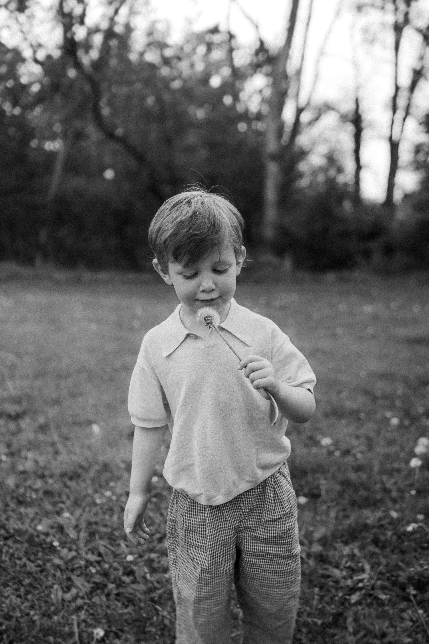 A black and white candid shot of a young child in a polo shirt standing in a wooded outdoor setting.