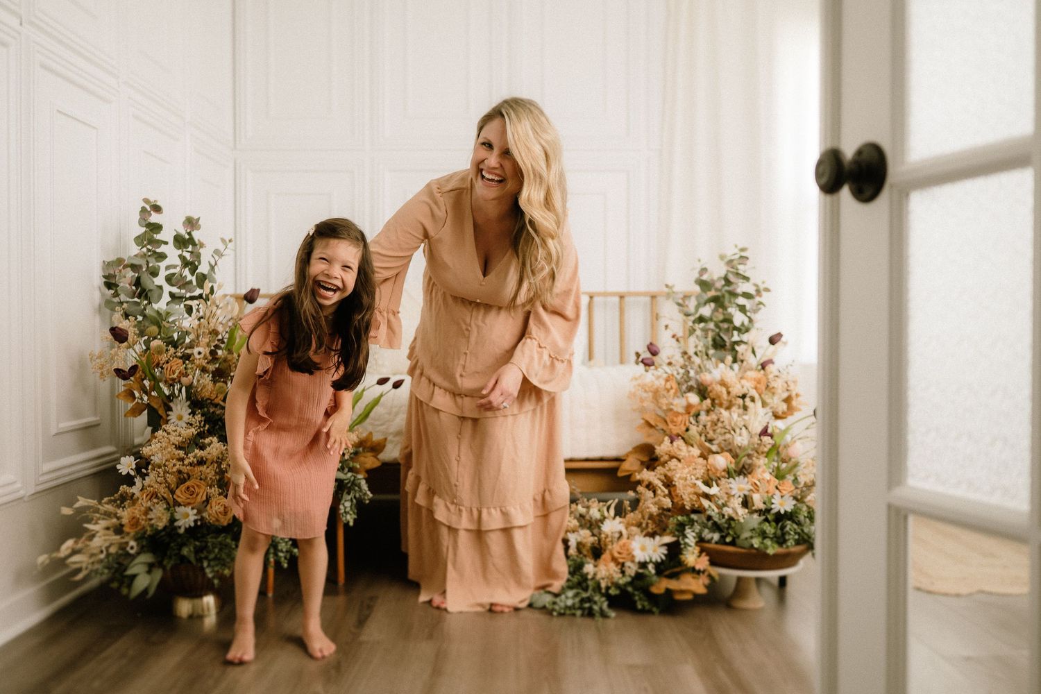 Two people wearing matching peach-colored ruffle dresses pose by decorated Christmas trees in a bright interior setting.