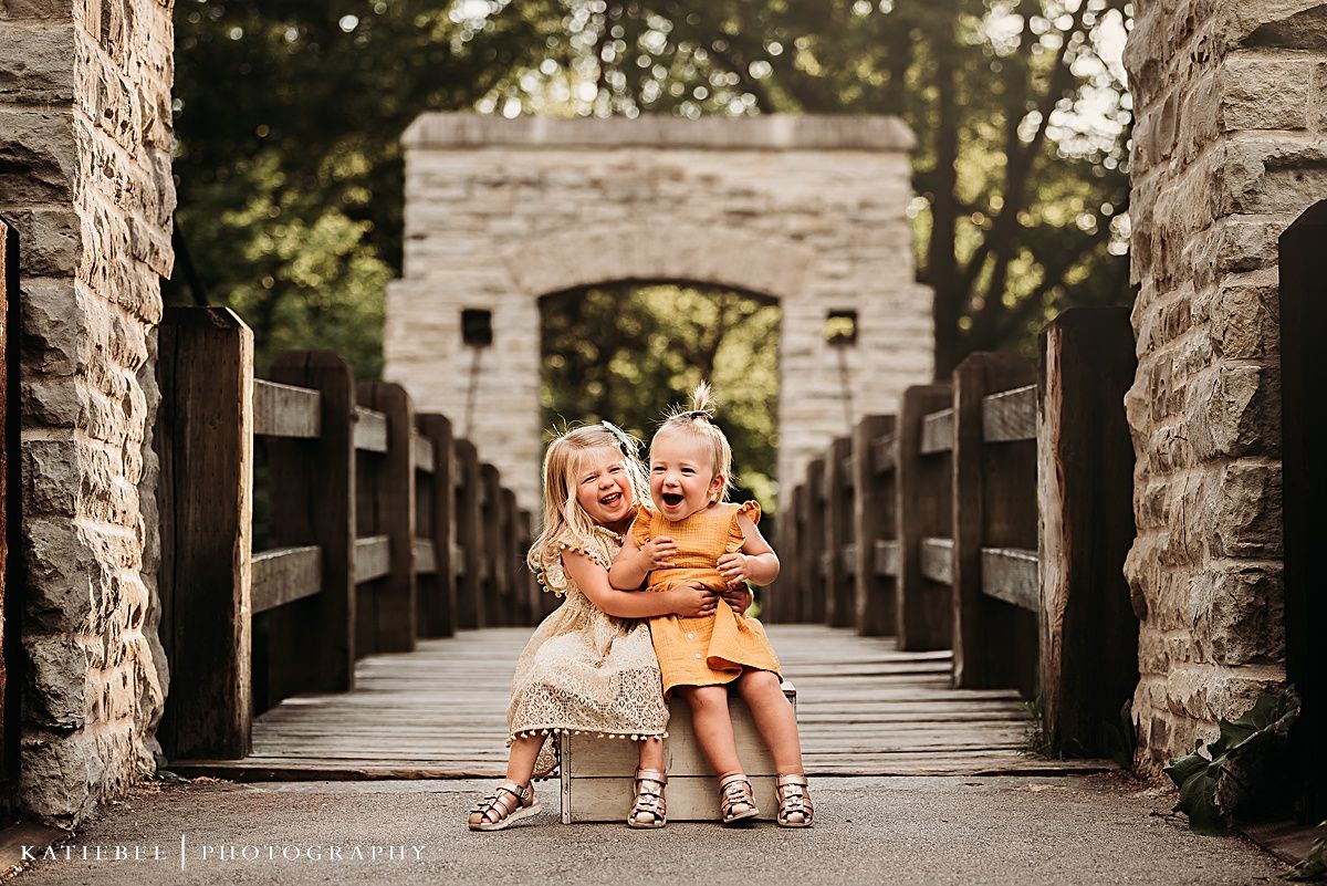 Two children sitting together on a wooden bench on a rustic bridge with stone archway.