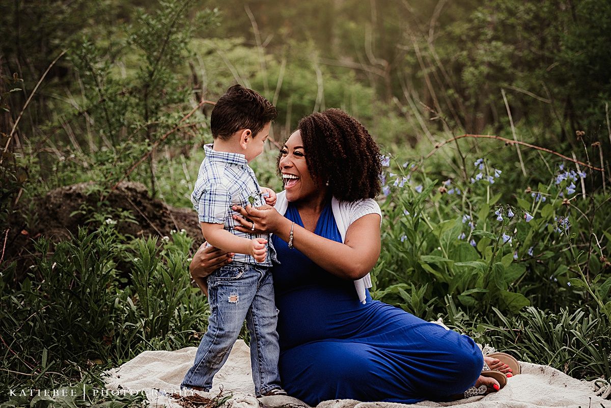 Mother and child share playful laughter while sitting outdoors on a blanket surrounded by greenery.