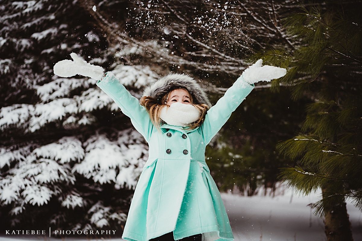 Person in mint coat with fur hood throwing arms up in snowy scene.
