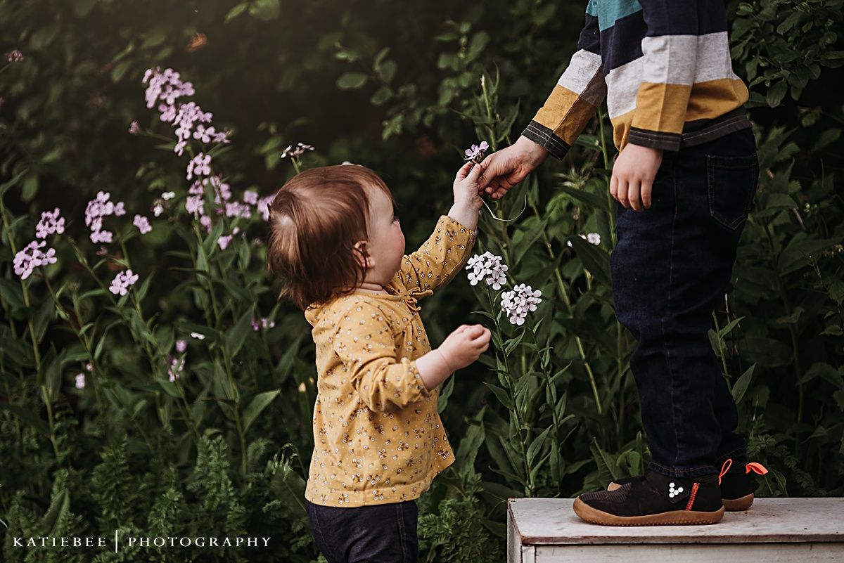 Small child in yellow shirt reaching up to smell purple wildflowers while standing on white box outdoors.