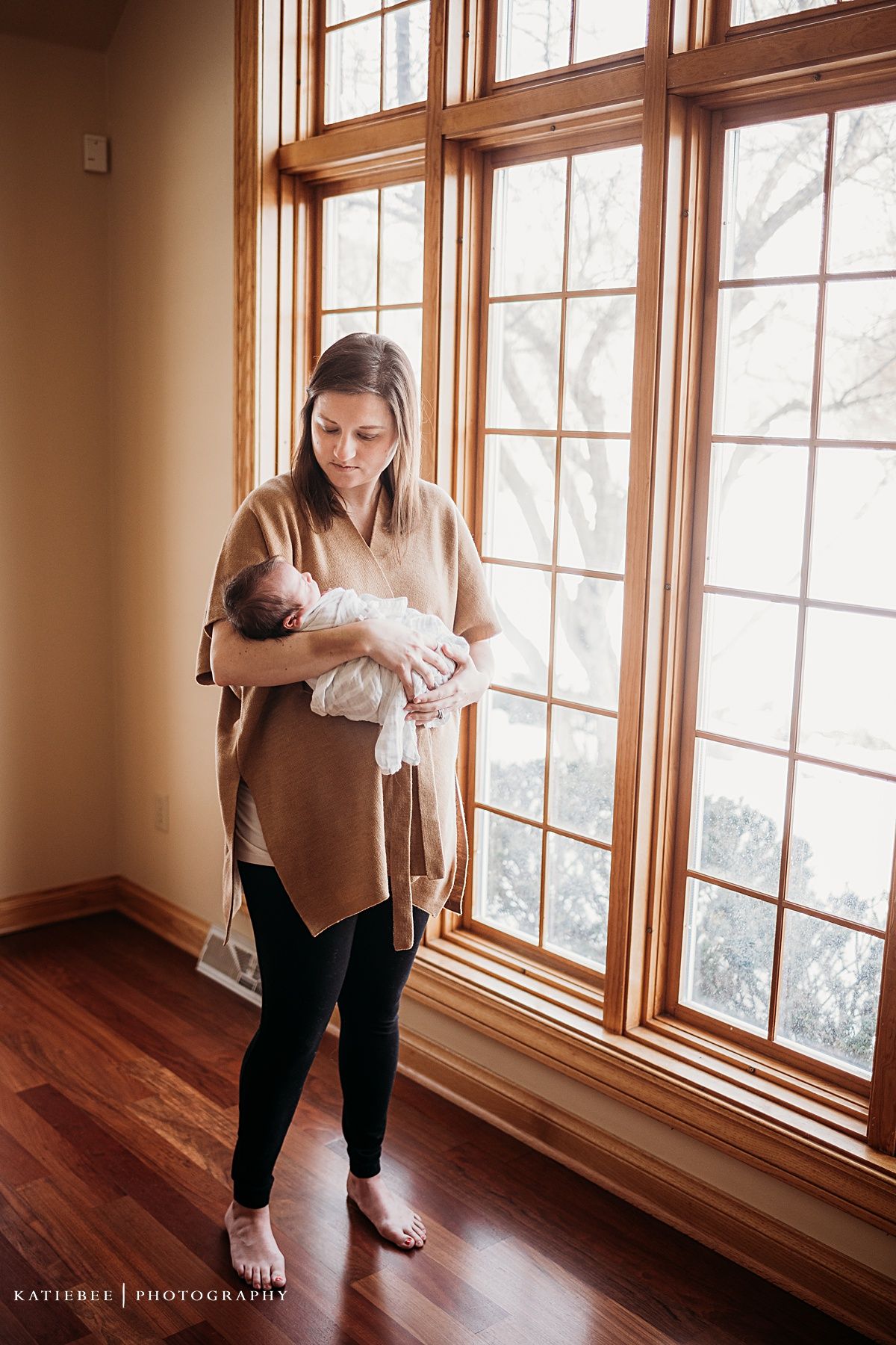 Mother holding newborn baby by large wooden framed windows in natural light.