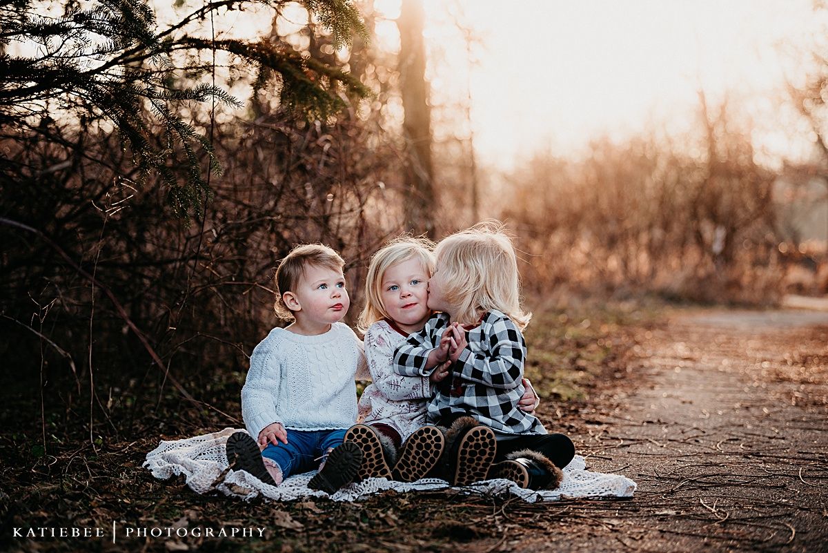 Three young children sit and interact playfully together on a nature trail at sunset.