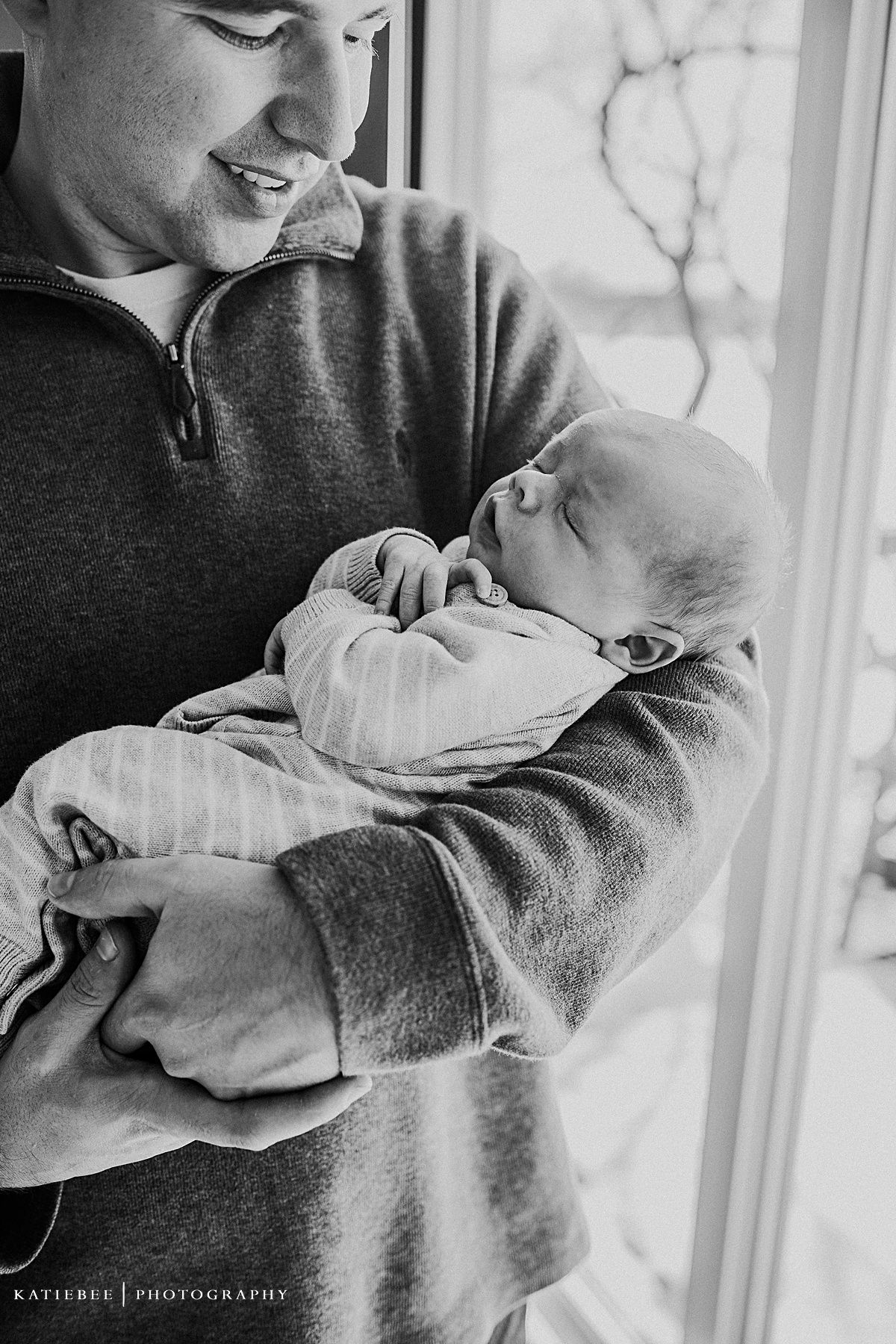Black and white portrait of parent cradling sleeping newborn by window.