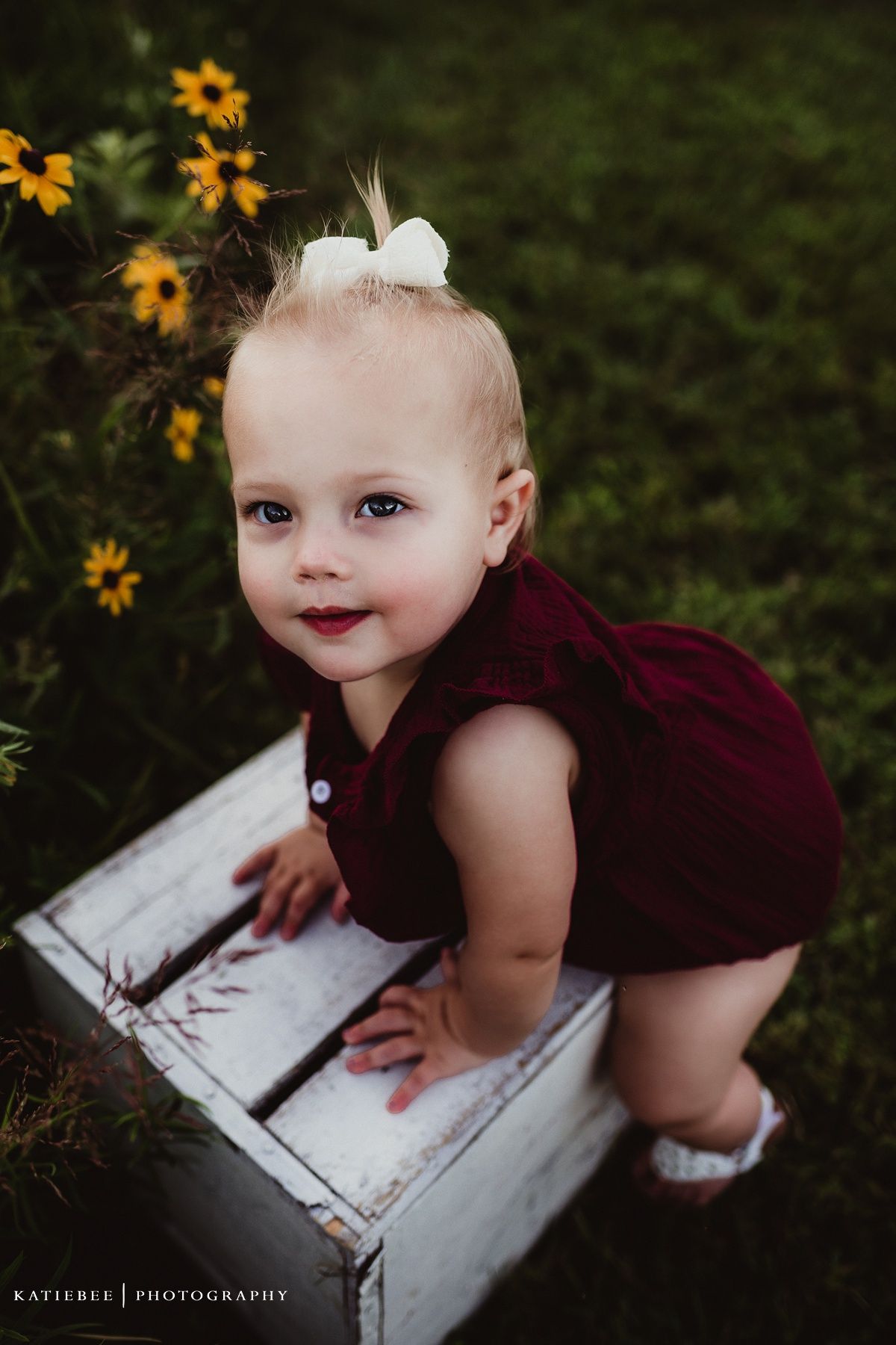 Smiling baby in burgundy dress leaning on white wooden box surrounded by yellow flowers.