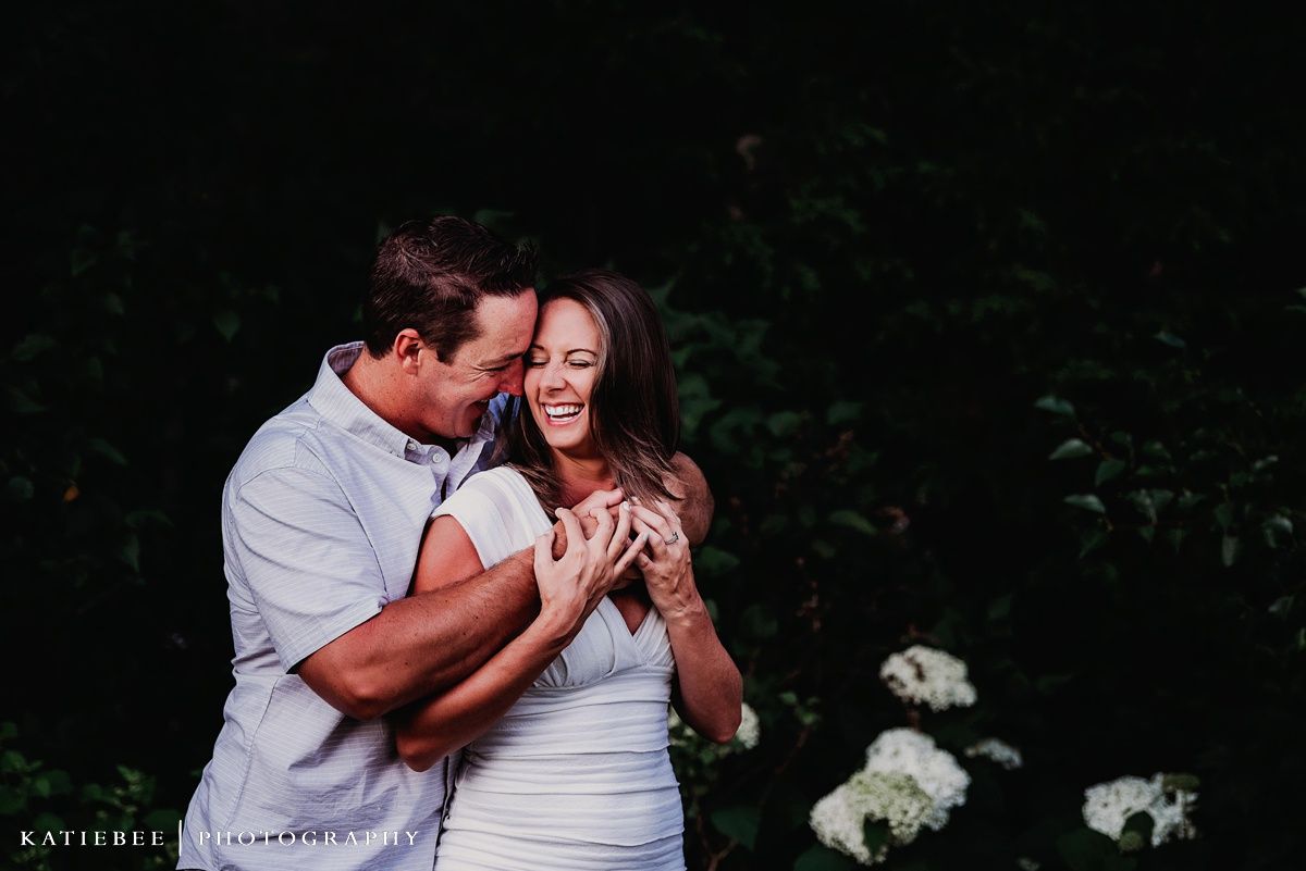 A couple embracing and laughing together outdoors near white flowers.