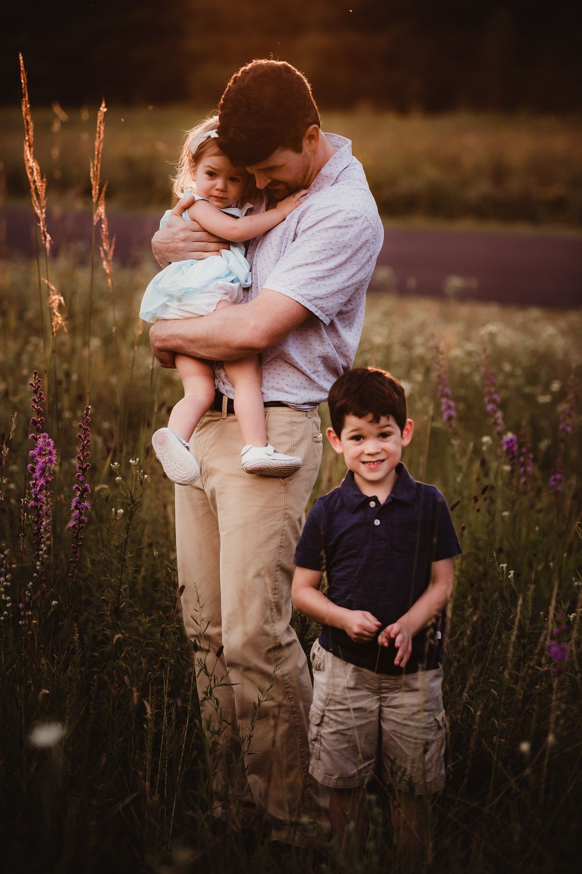 Tender family moment captured in golden sunlit field with purple wildflowers at sunset.