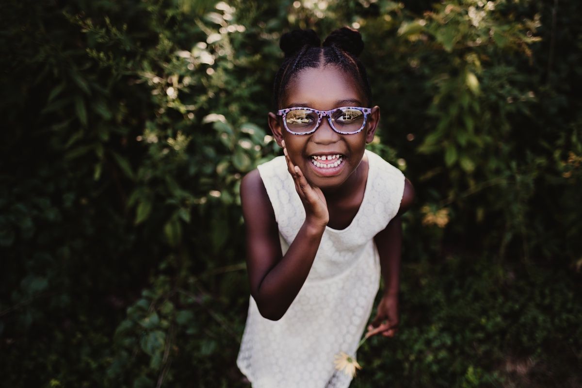 Child wearing glasses and white dress smiling joyfully in an outdoor garden setting.