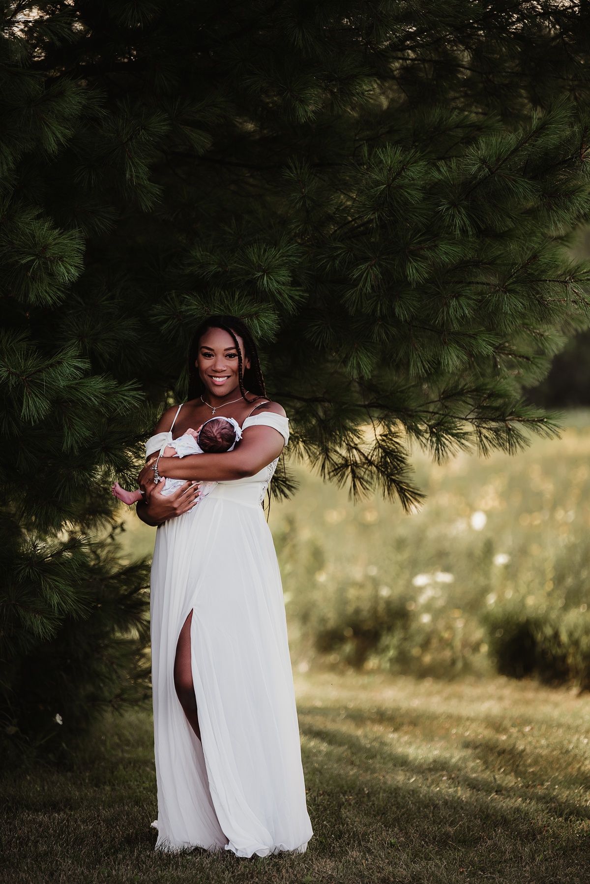 An intimate portrait of someone in a white dress holding a baby outdoors under pine trees.