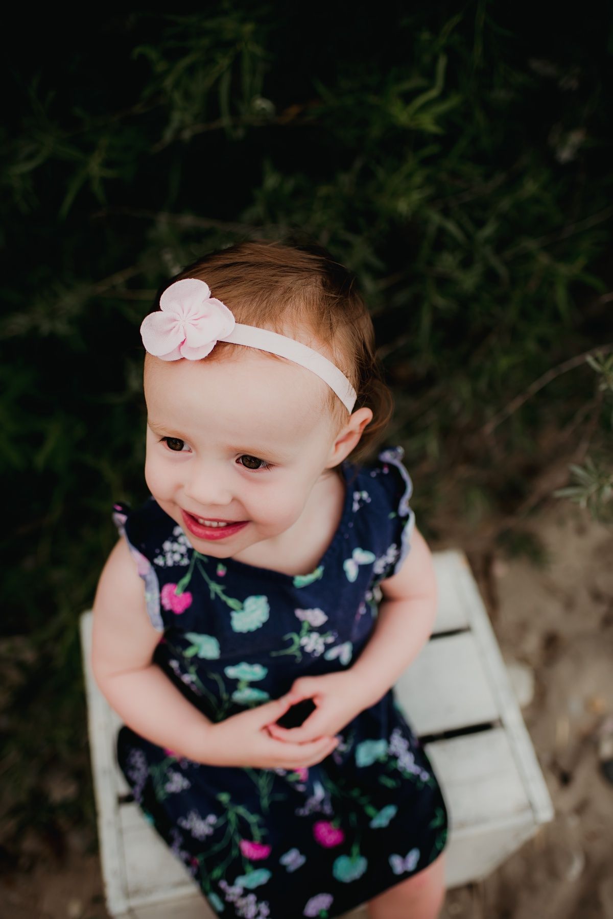 A toddler in a floral dress and white headband smiles while sitting on a wooden surface.