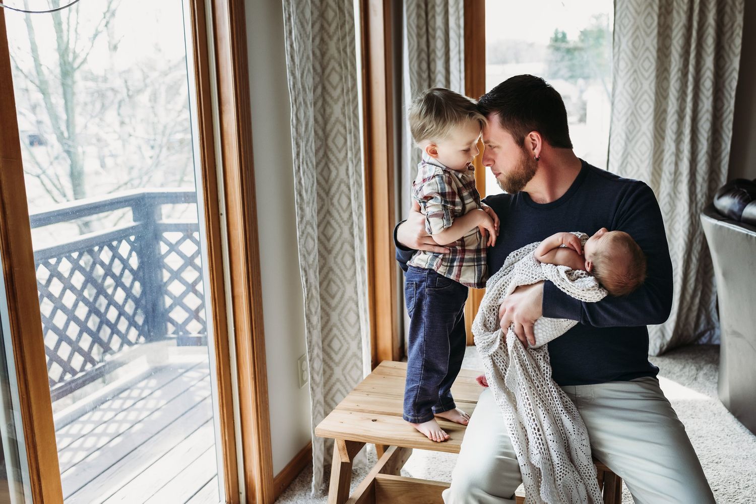 Parent and toddler stand together by a window while holding a swaddled newborn.