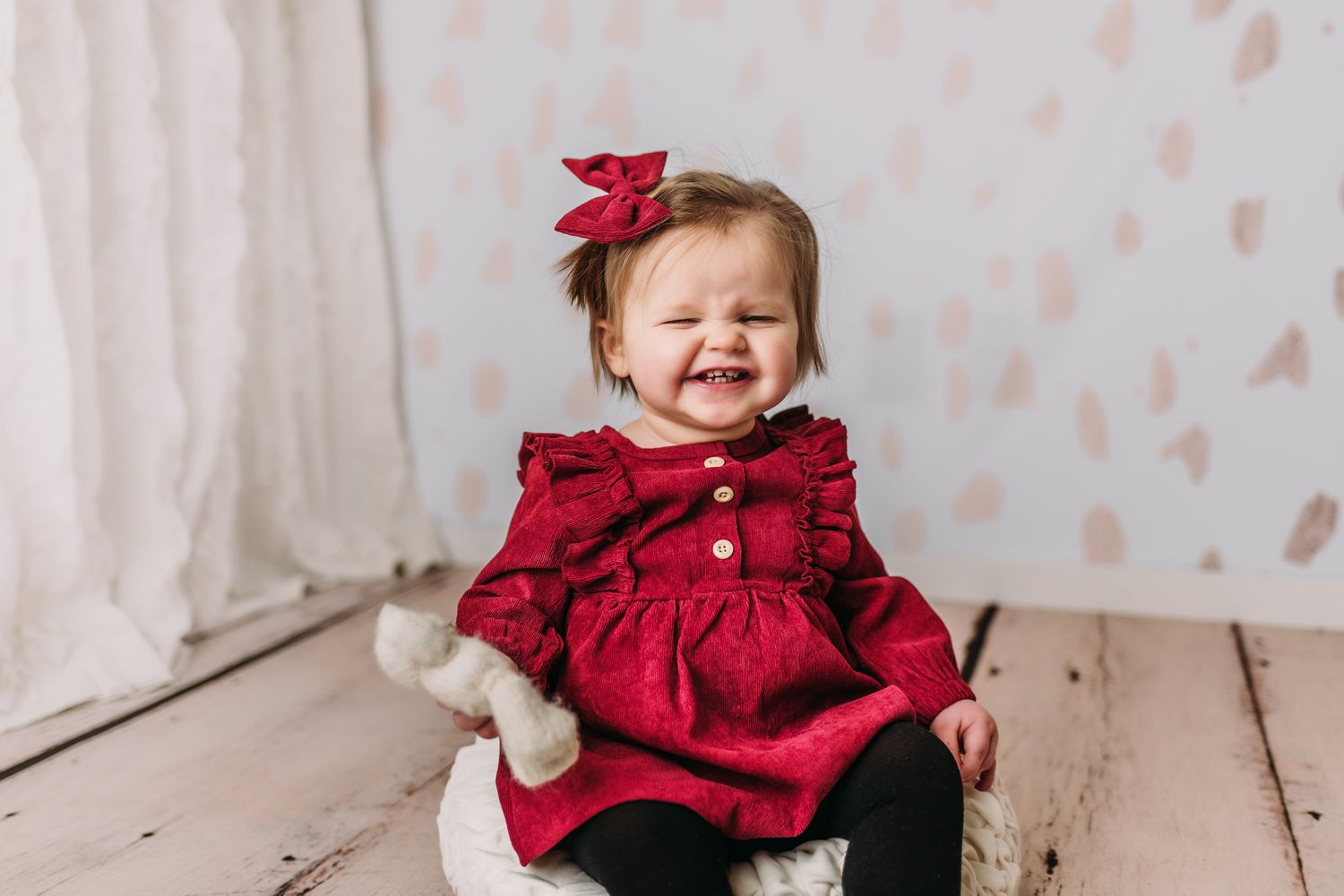 Baby wearing festive red dress with matching bow sits smiling on white wooden floor.