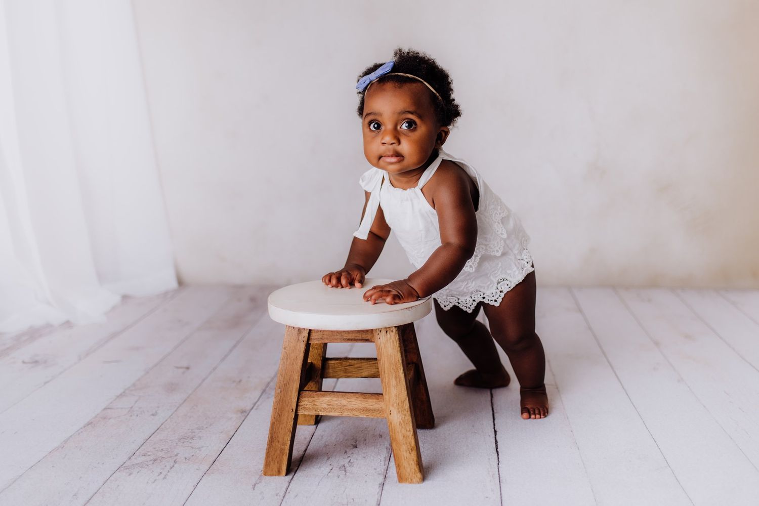 A toddler in white clothing leans against a wooden stool on a light wooden floor.