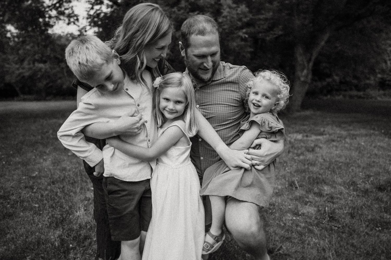 Black and white intimate family portrait taken outdoors against trees.