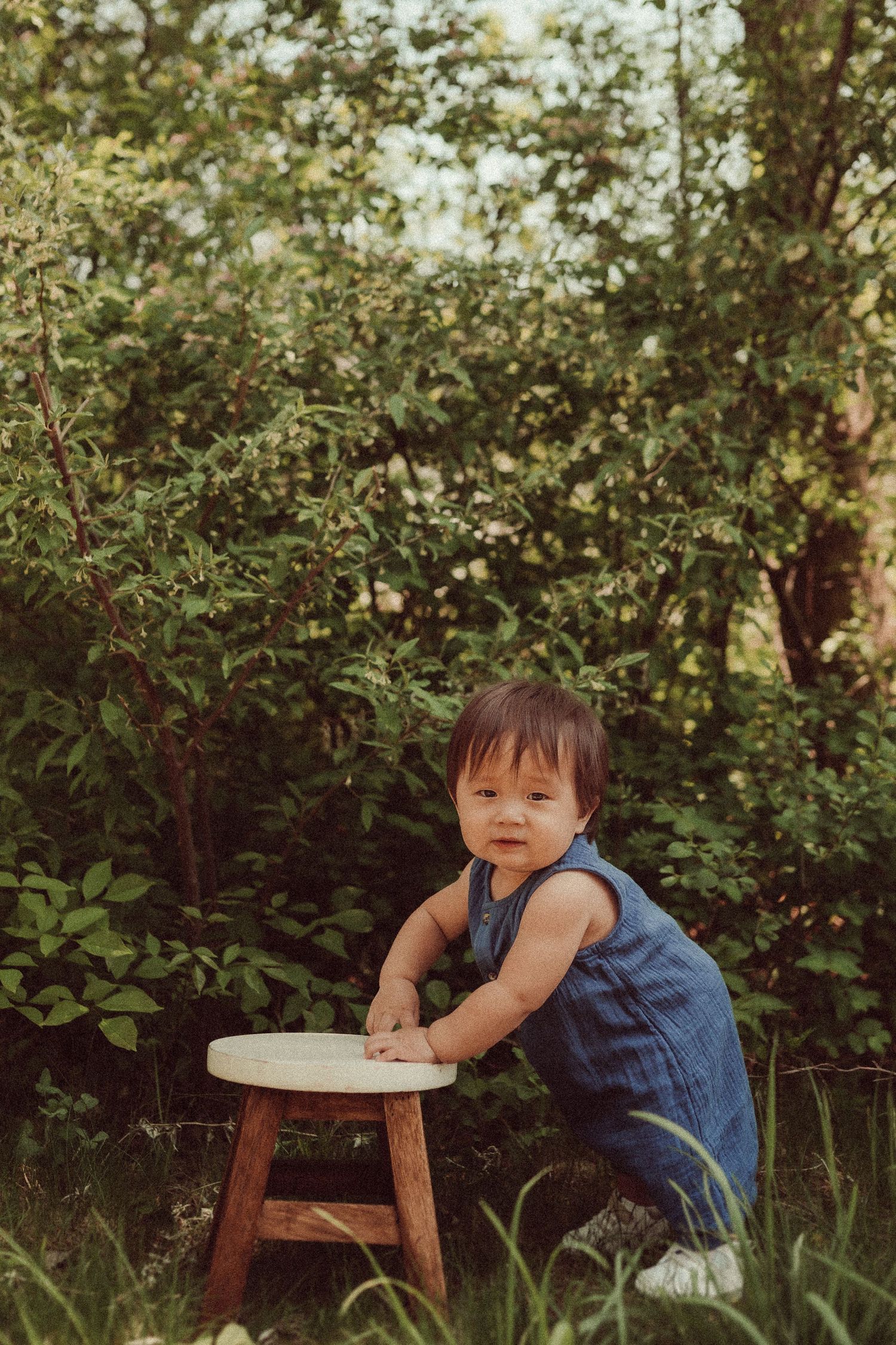 A toddler in denim overalls plays with a wooden stool outdoors among green foliage.