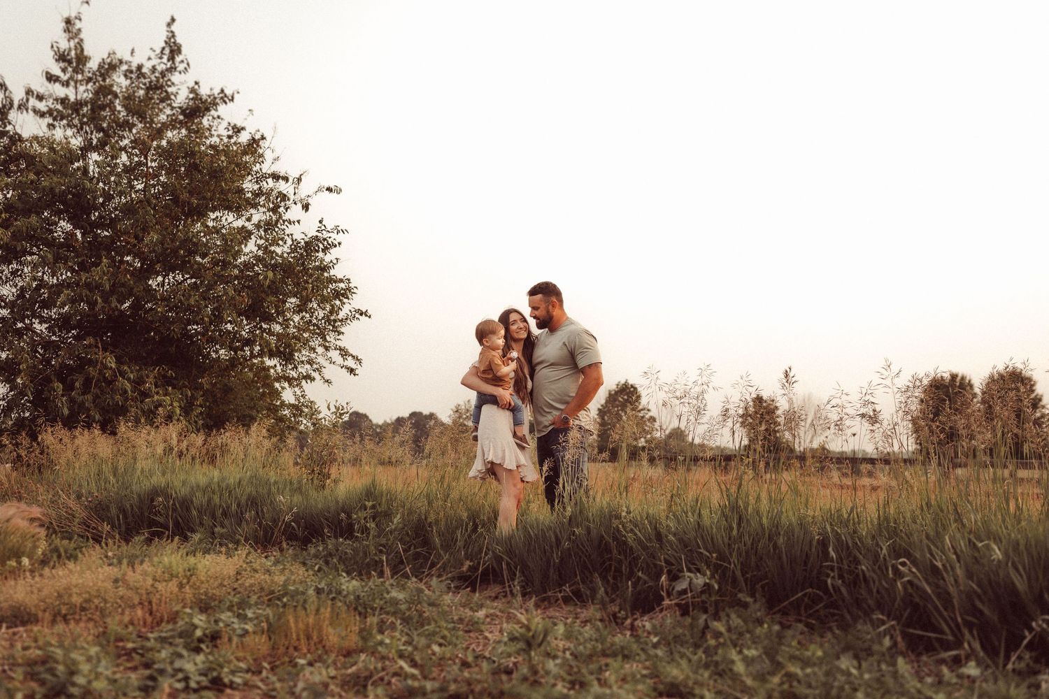 A loving family portrait captured at golden hour in a rustic field with tall grass and trees in the background.