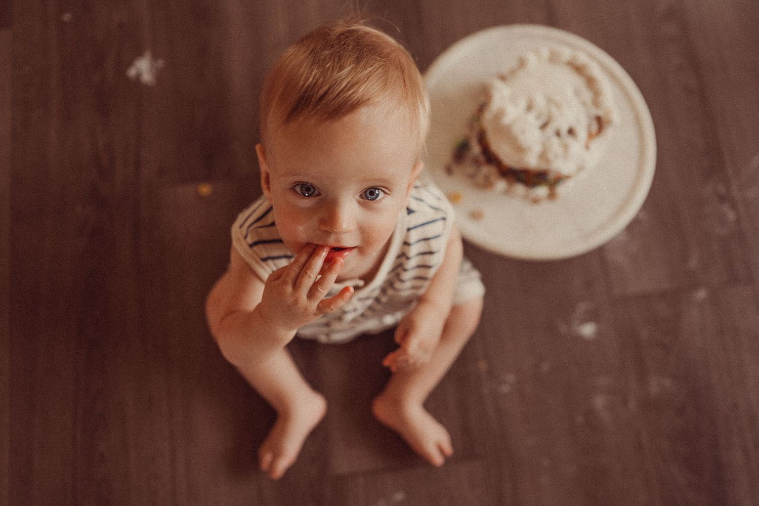 Overhead view of toddler in striped outfit enjoying cake smash photo session on hardwood floor.