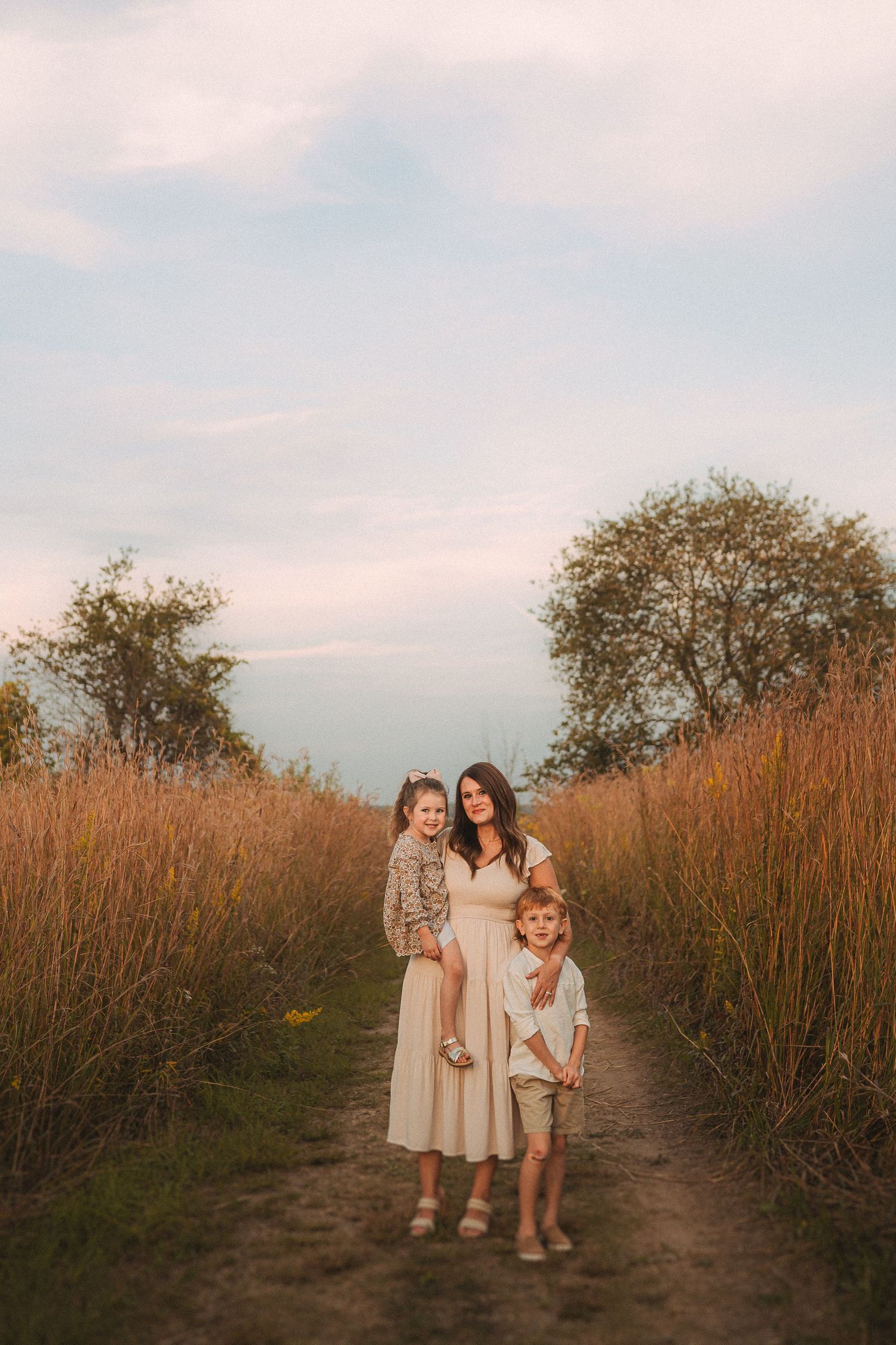 Three figures in light dresses standing together on nature path during golden hour.