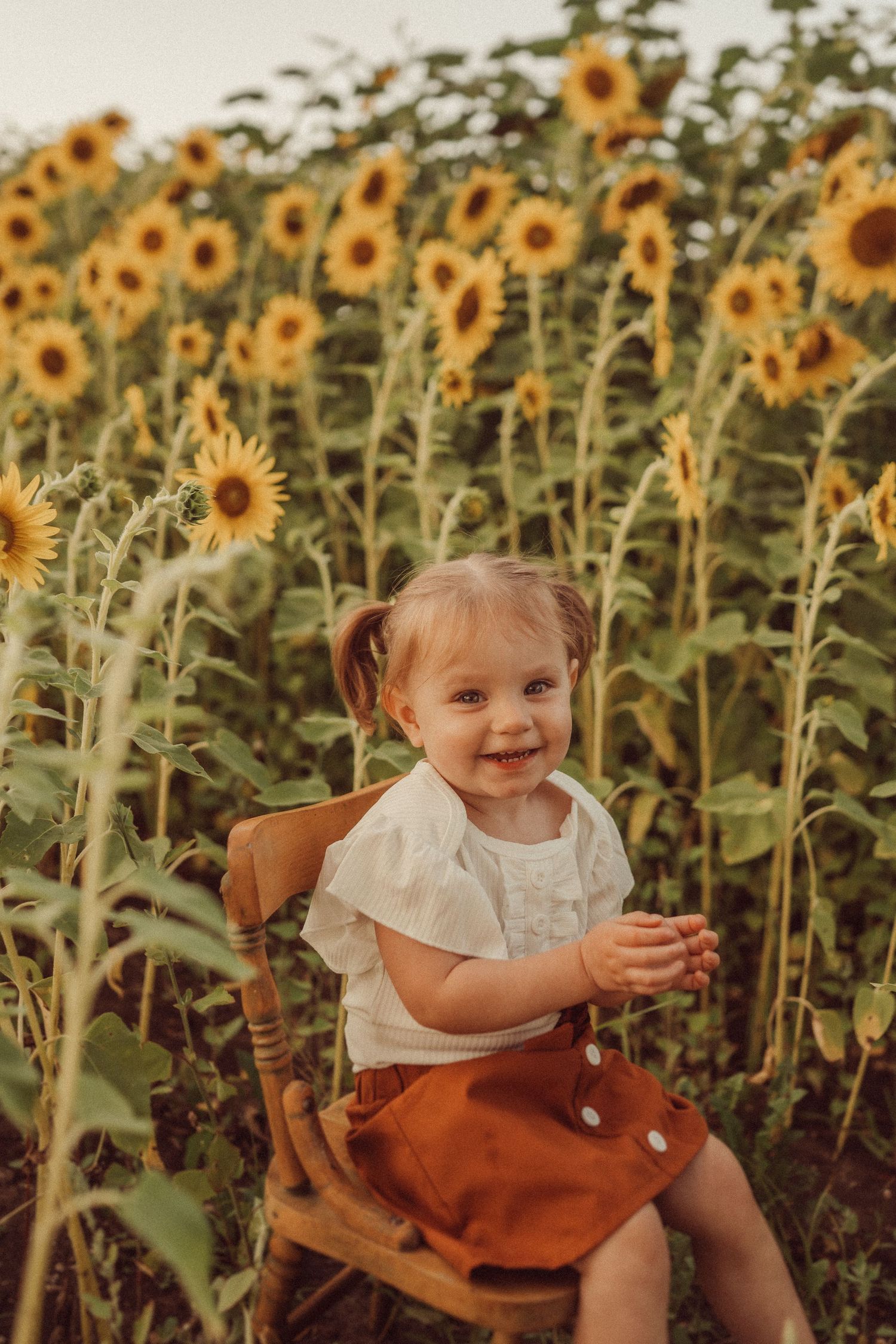 Young child sits smiling in wooden chair surrounded by tall sunflowers.