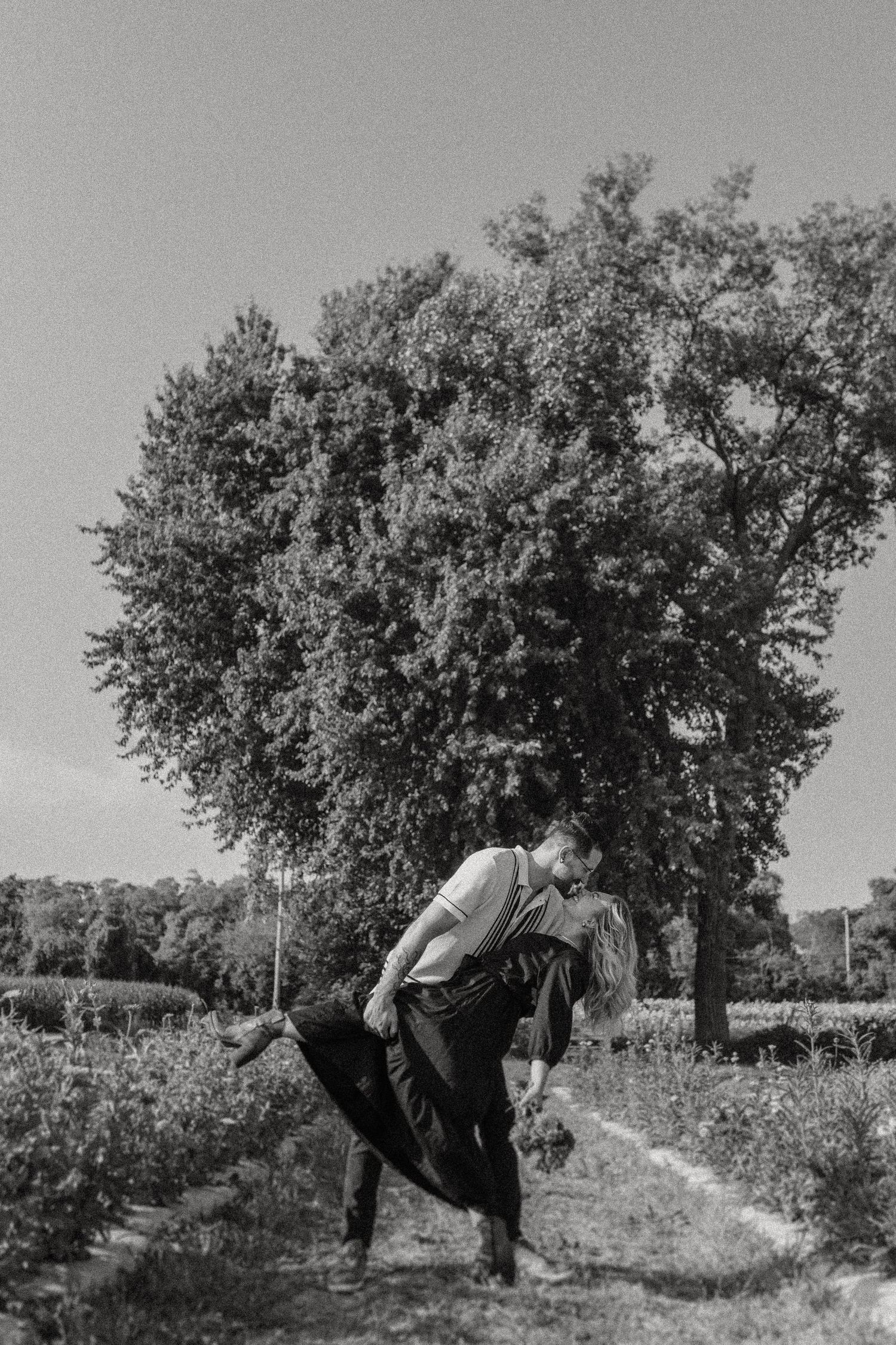 Black and white photo of a couple in a playful dance pose outdoors.