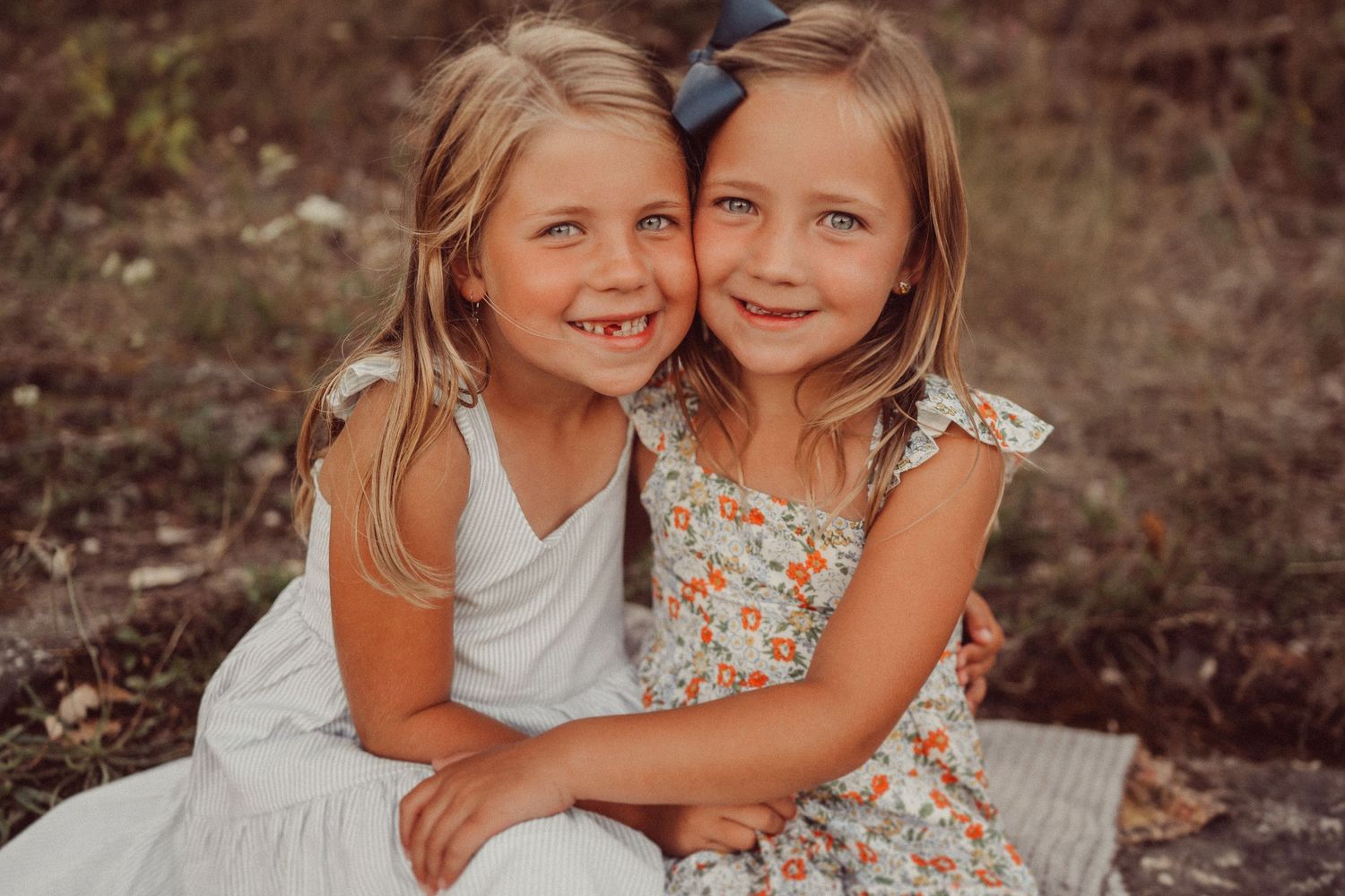 Two children embrace while sitting together outdoors in complementary white and floral dresses.