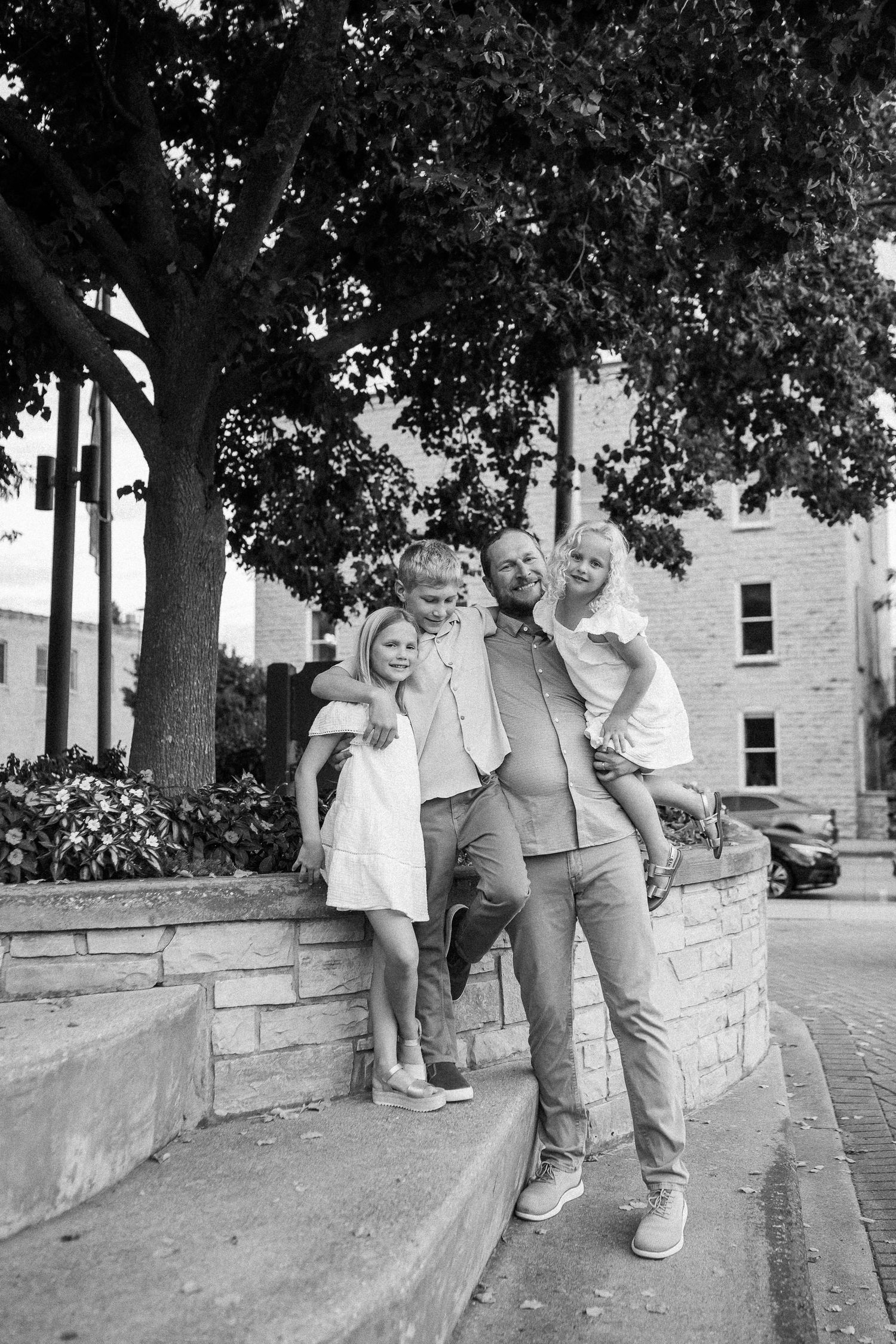 Black and white photo of people sitting and standing along a stone retaining wall under trees.
