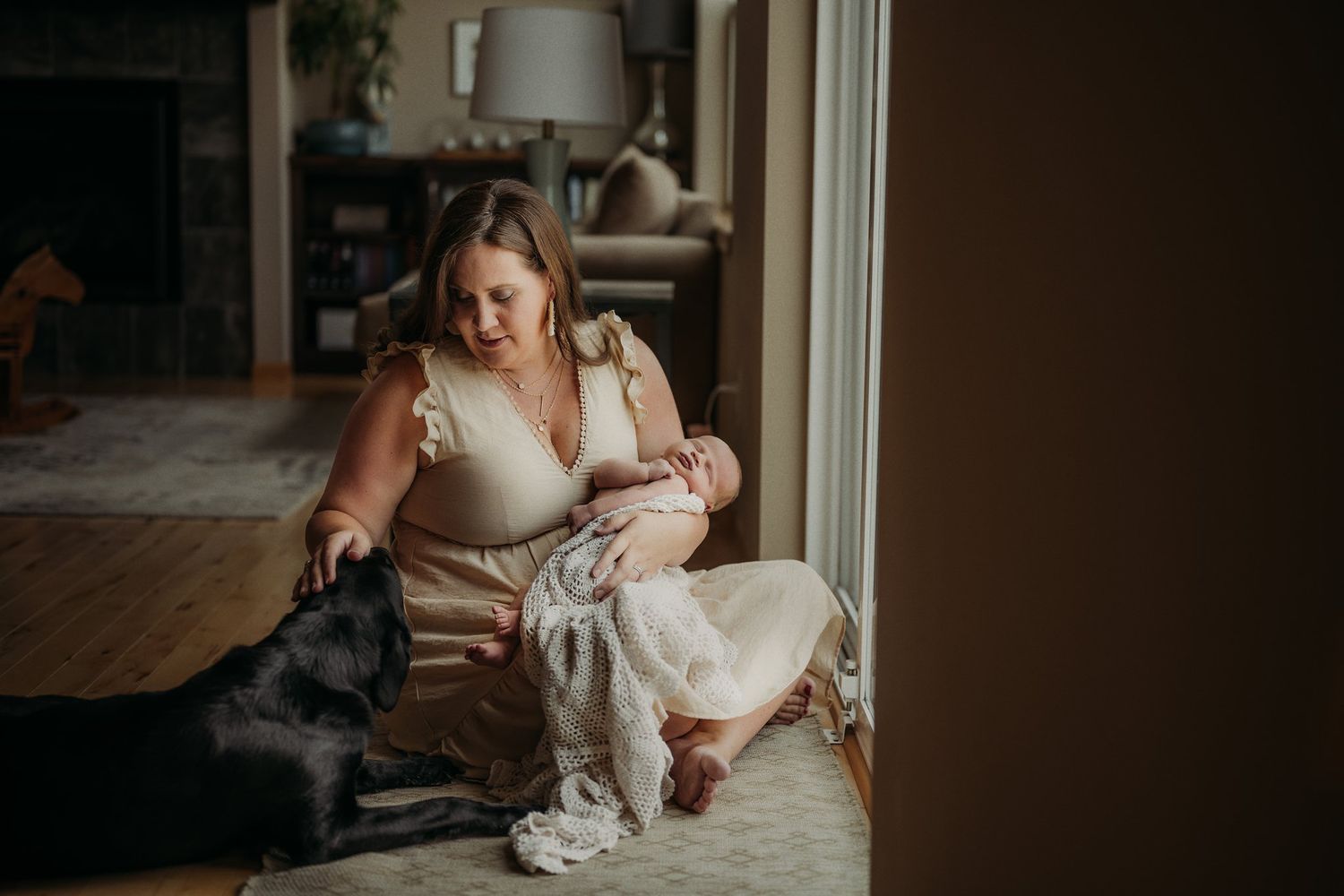 A family sits by a window at home with their black dog and infant on a cozy blanket.
