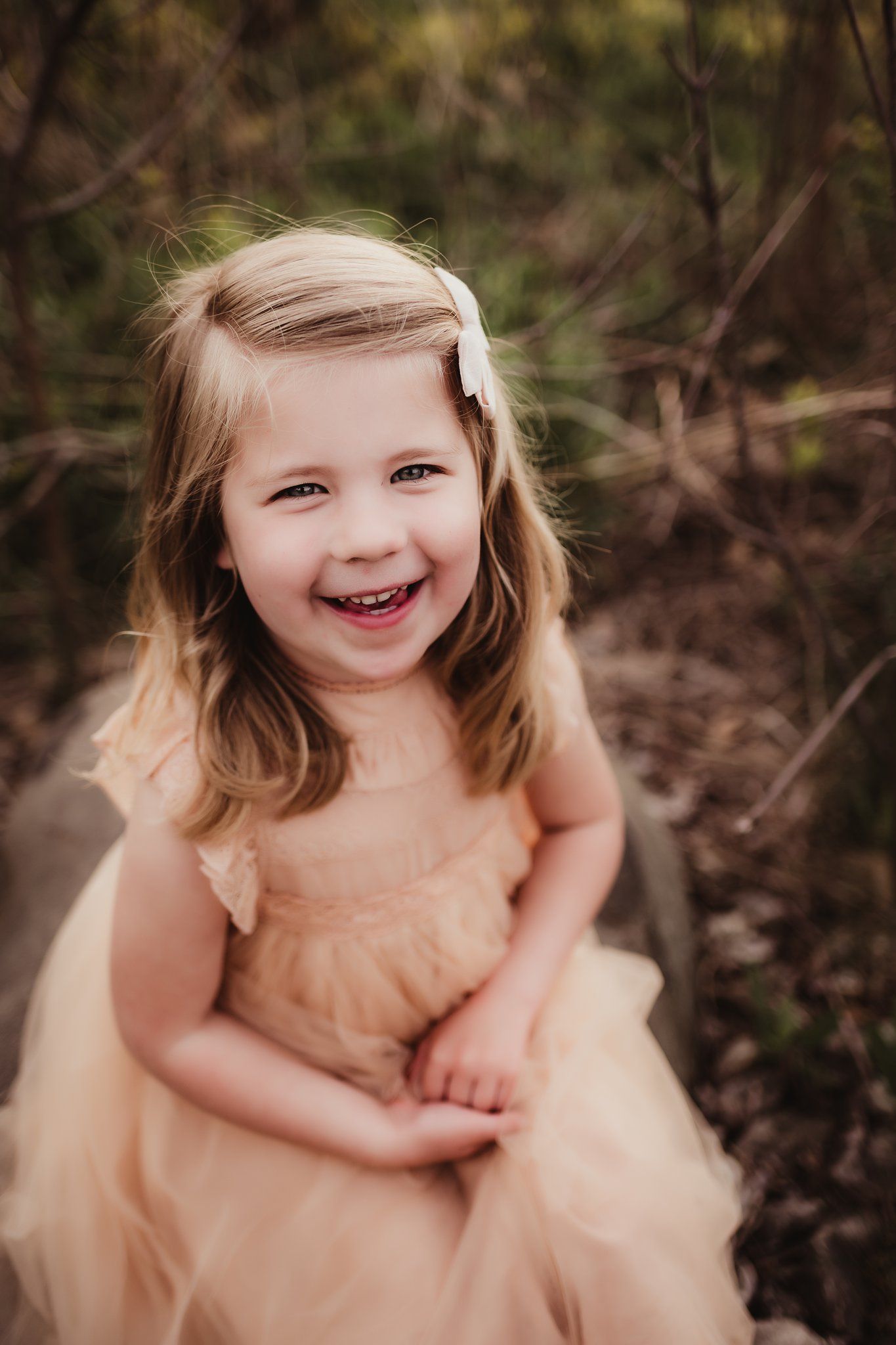 A child wearing a peachy dress smiles brightly against a natural backdrop.