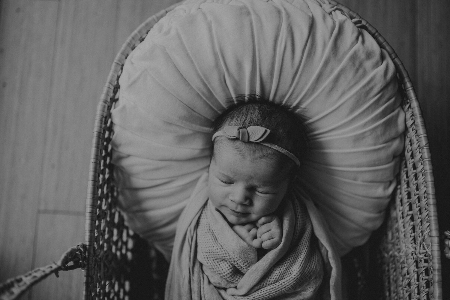 Newborn baby swaddled in beige wrap sleeping peacefully on round tufted pillow in black and white photograph.