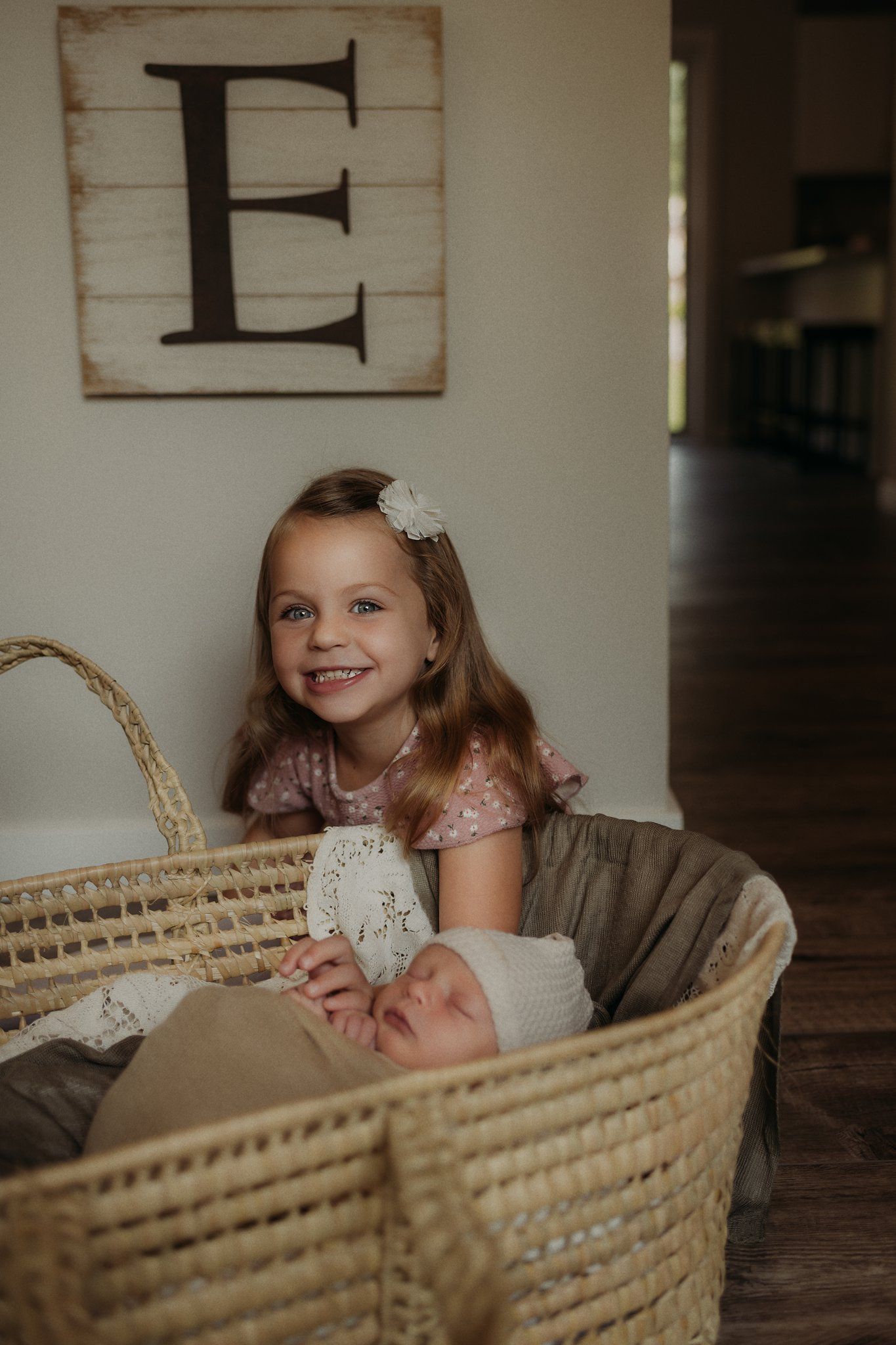 A young child peeks over a woven basket containing an infant under a decorative letter E wall hanging.