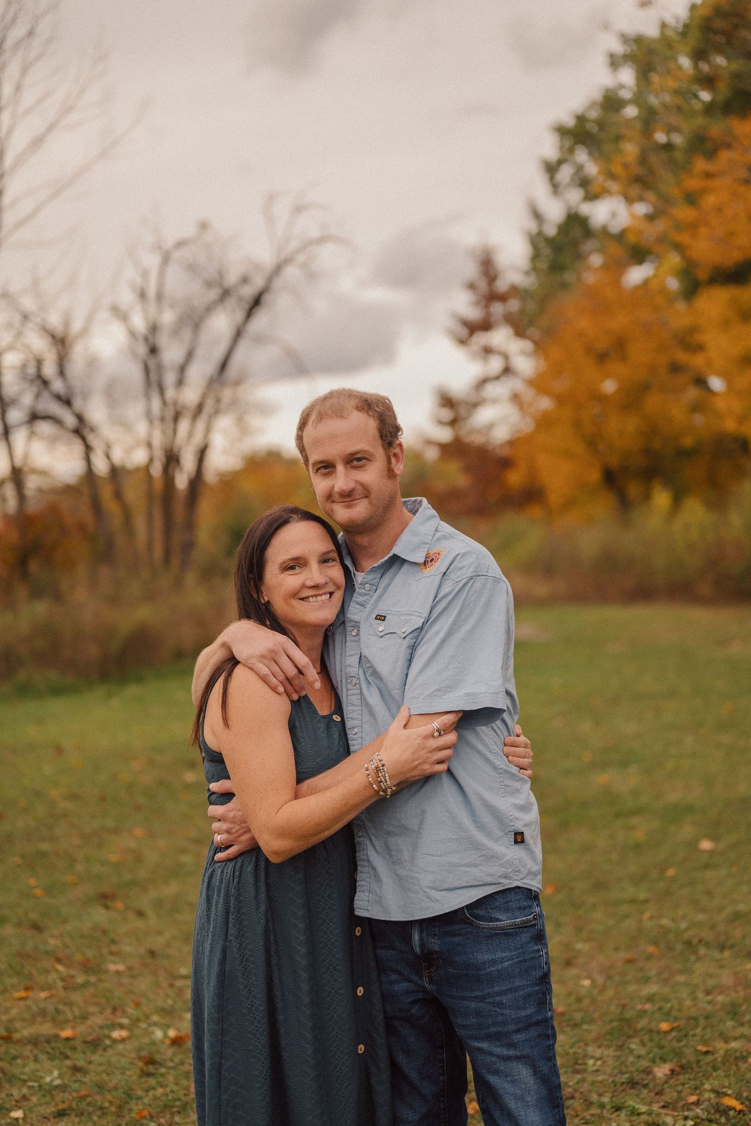Couple embracing in a field surrounded by vibrant fall colors and trees.