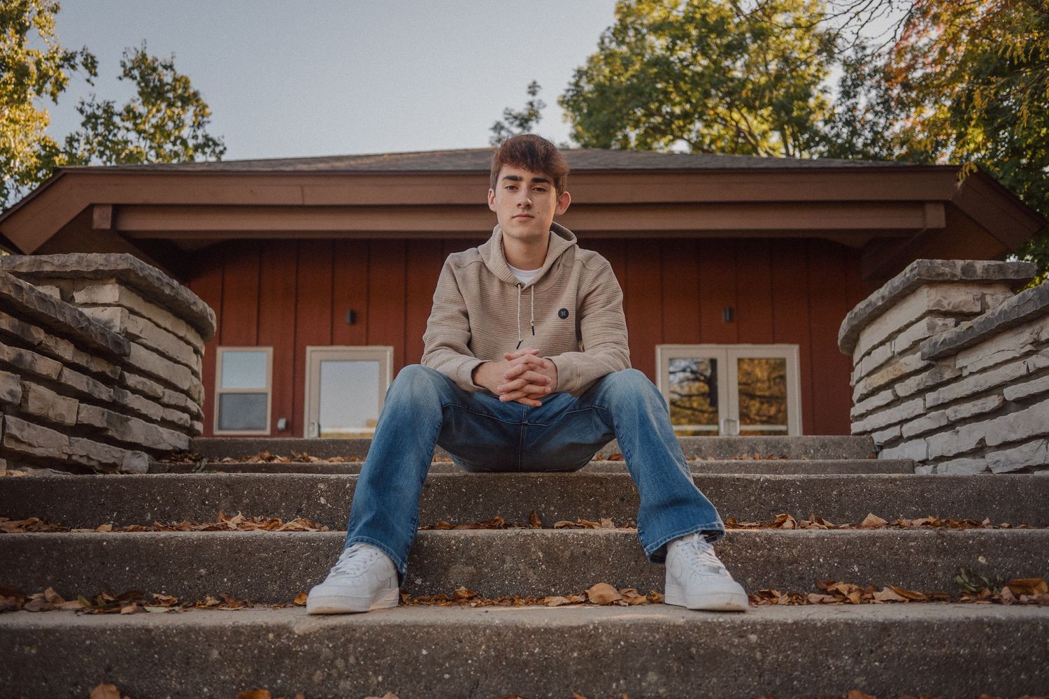 A person in casual attire sits on stone steps surrounded by autumn leaves on a sunny day.