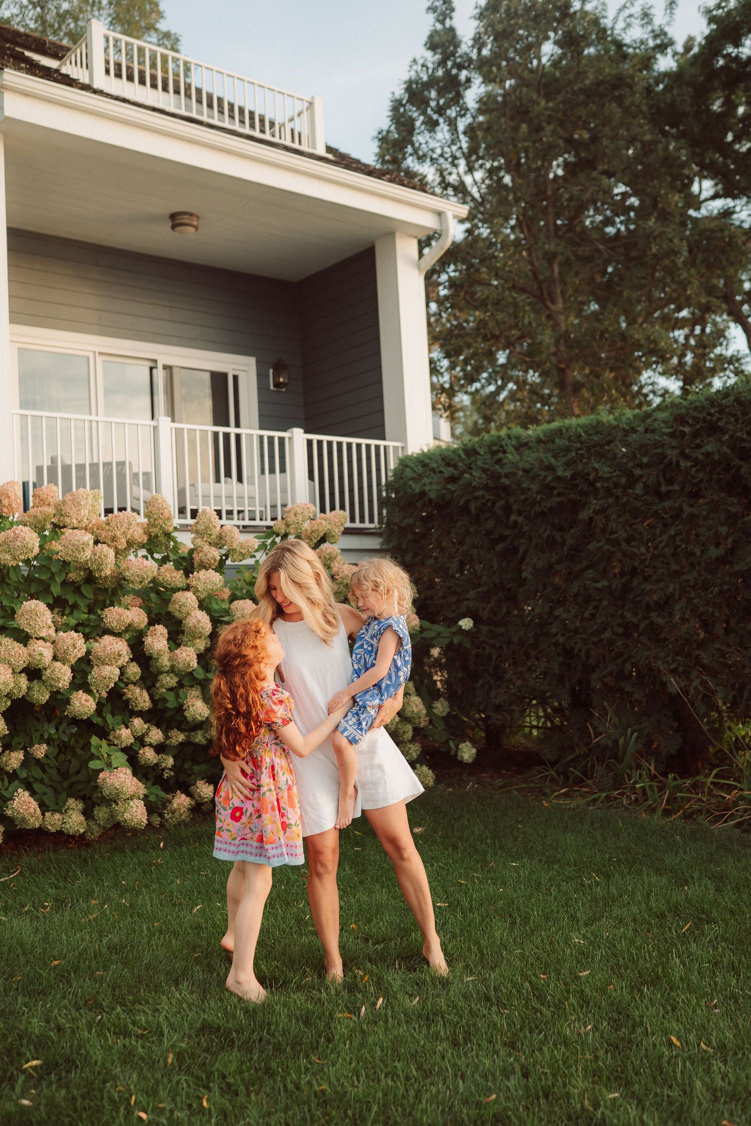 Three people play together on a lush green lawn in front of a house with white railings and blooming hydrangeas.