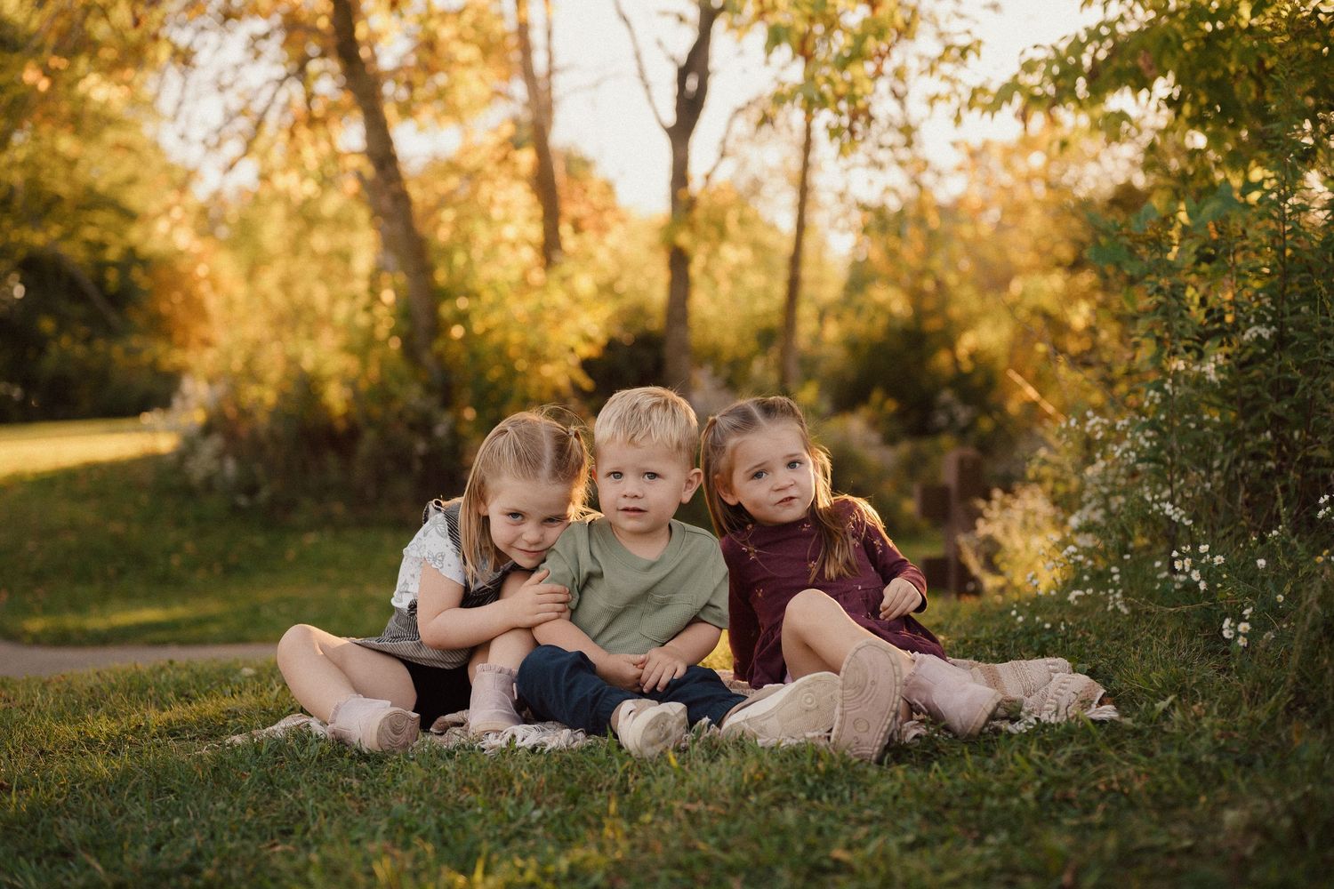 Family lying together on autumn grass during golden hour with sunlit trees in background.