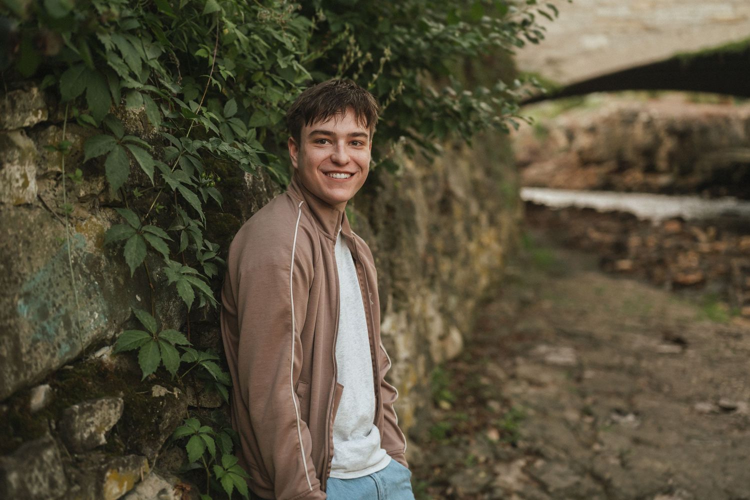 A person in a brown jacket stands near stone walls and greenery in an outdoor setting.