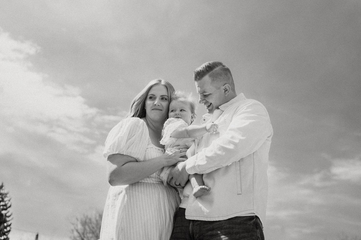 A black and white photo of parents lovingly holding their baby under a dramatic cloudy sky.