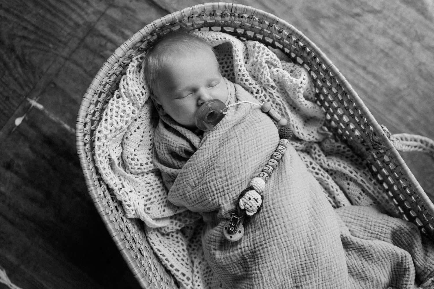 Black and white photo of a sleeping newborn baby wrapped in a knit blanket inside a wicker Moses basket.