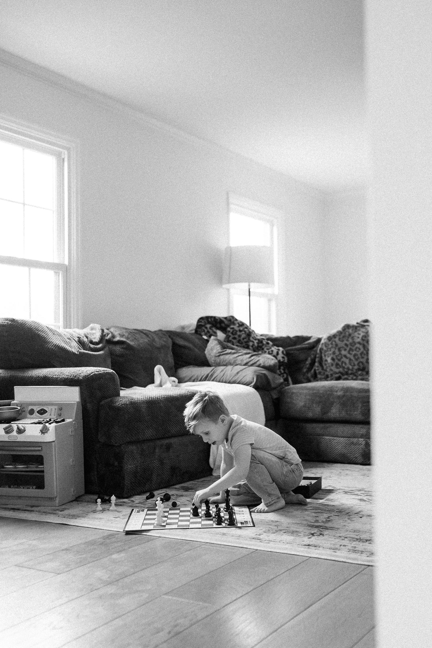 Black and white photo of a young boy playing chess alone on a living room floor near a couch.