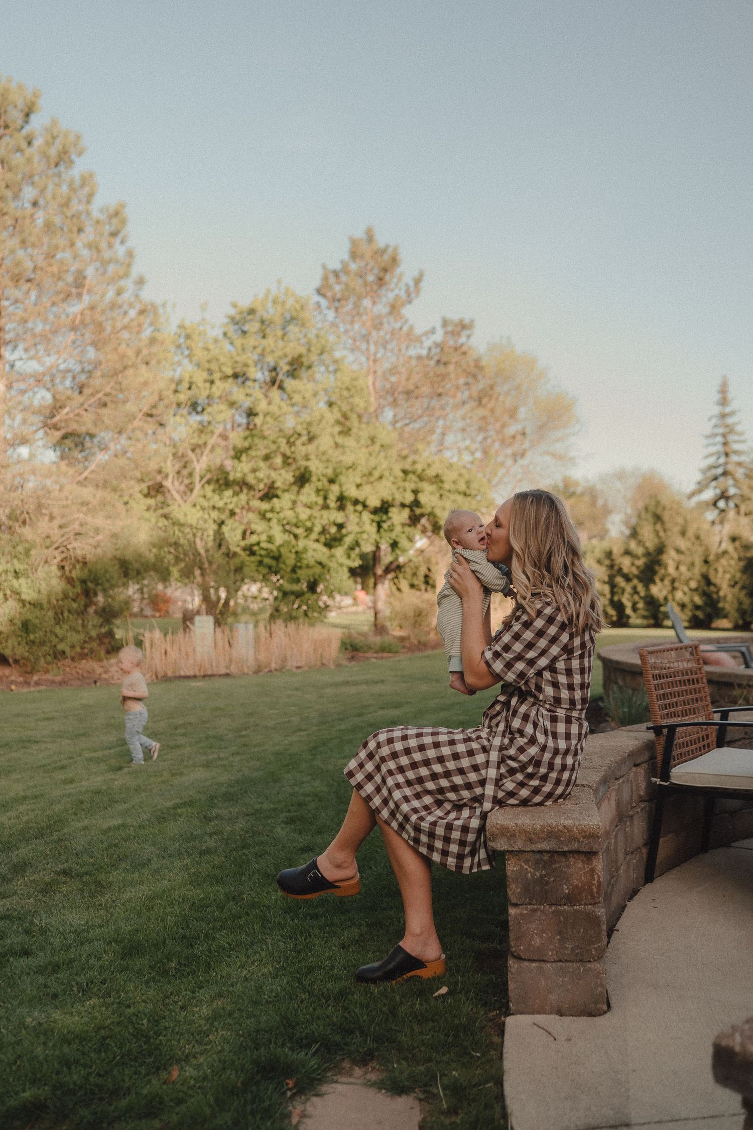 Mother in gingham dress sitting outdoors kissing baby while toddler plays in green backyard.
