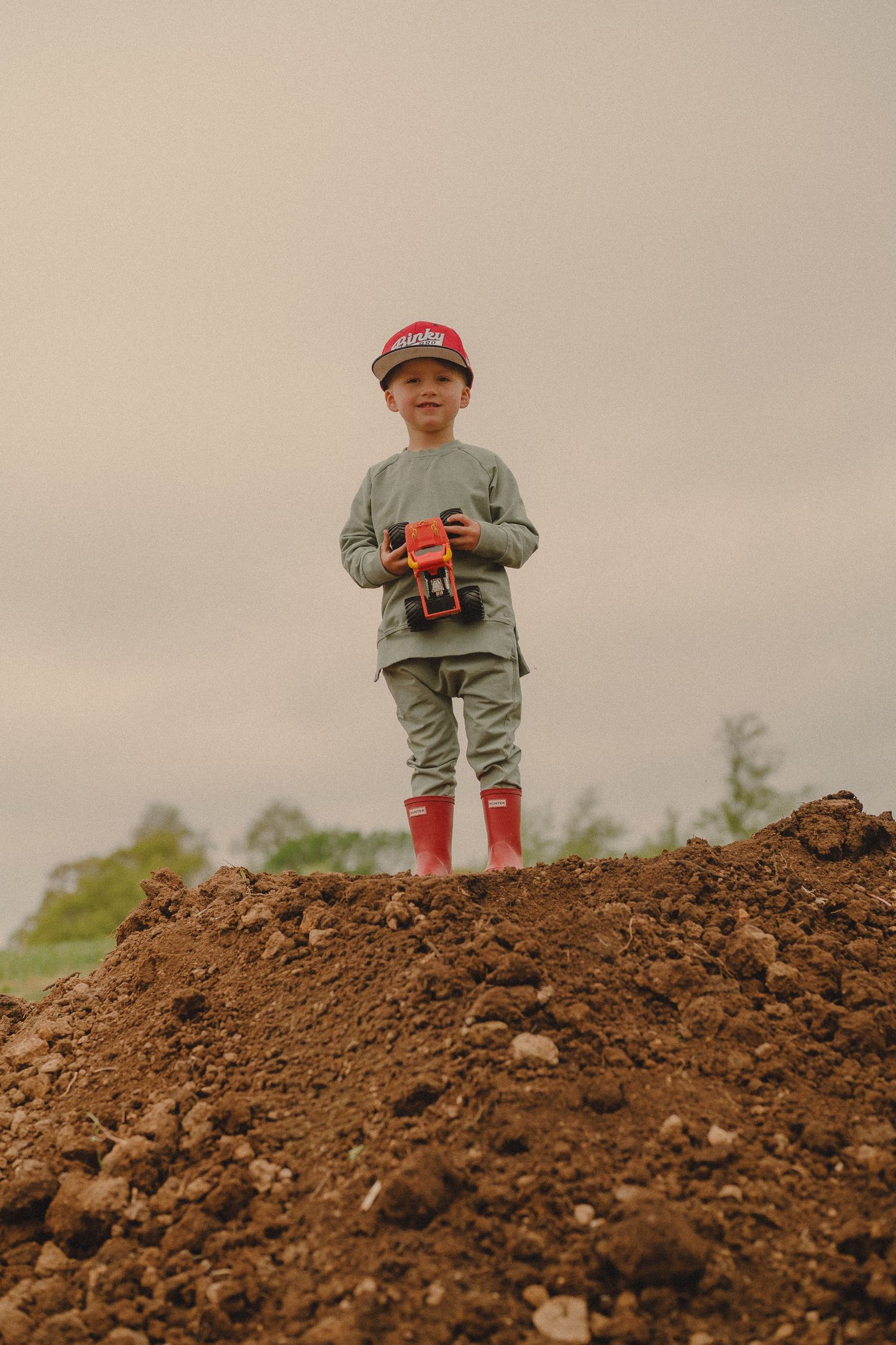 Young boy standing on dirt mound holding red toy truck wearing red boots and cap.