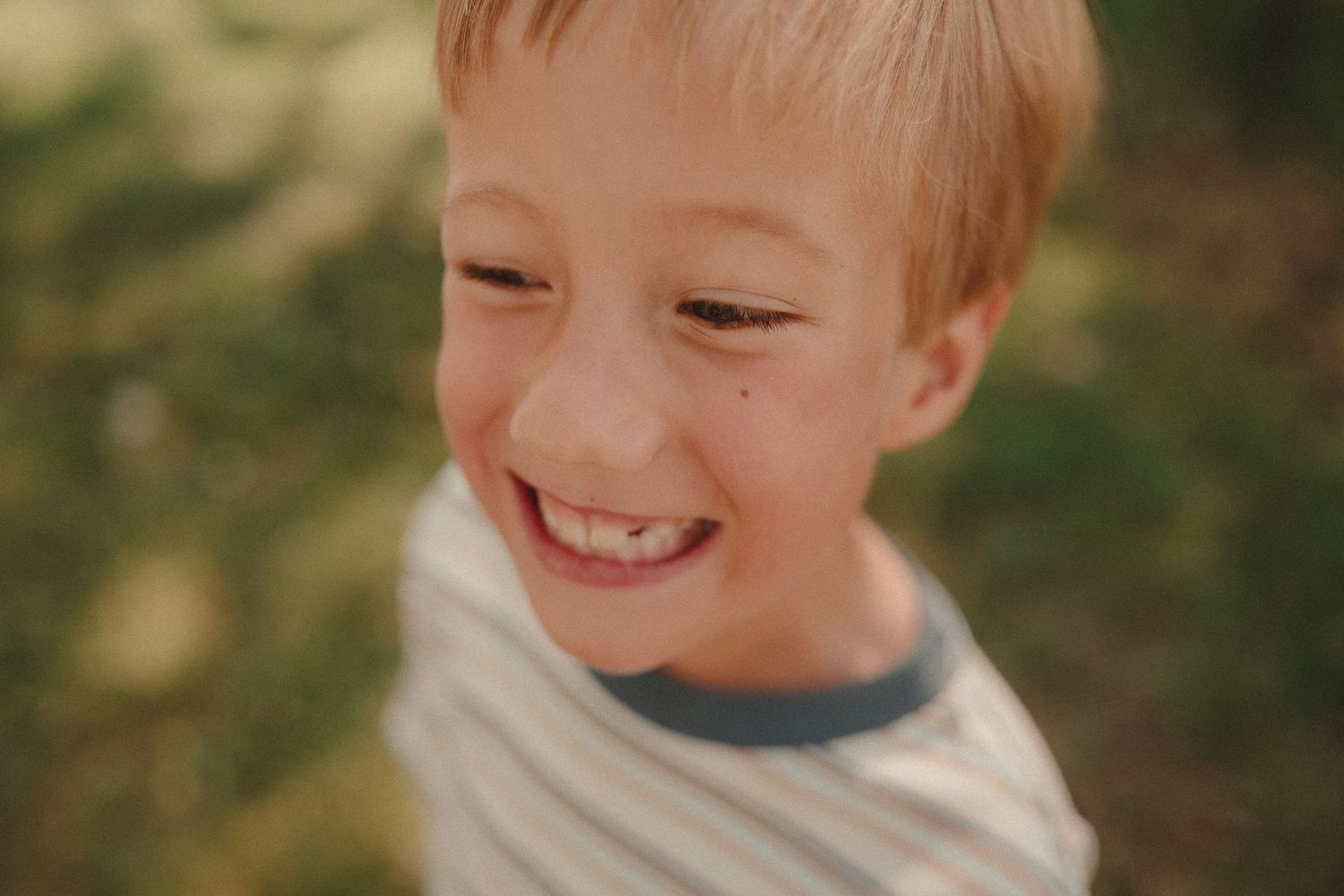 A freckled blond boy grins joyfully outdoors in warm natural light wearing a striped shirt.