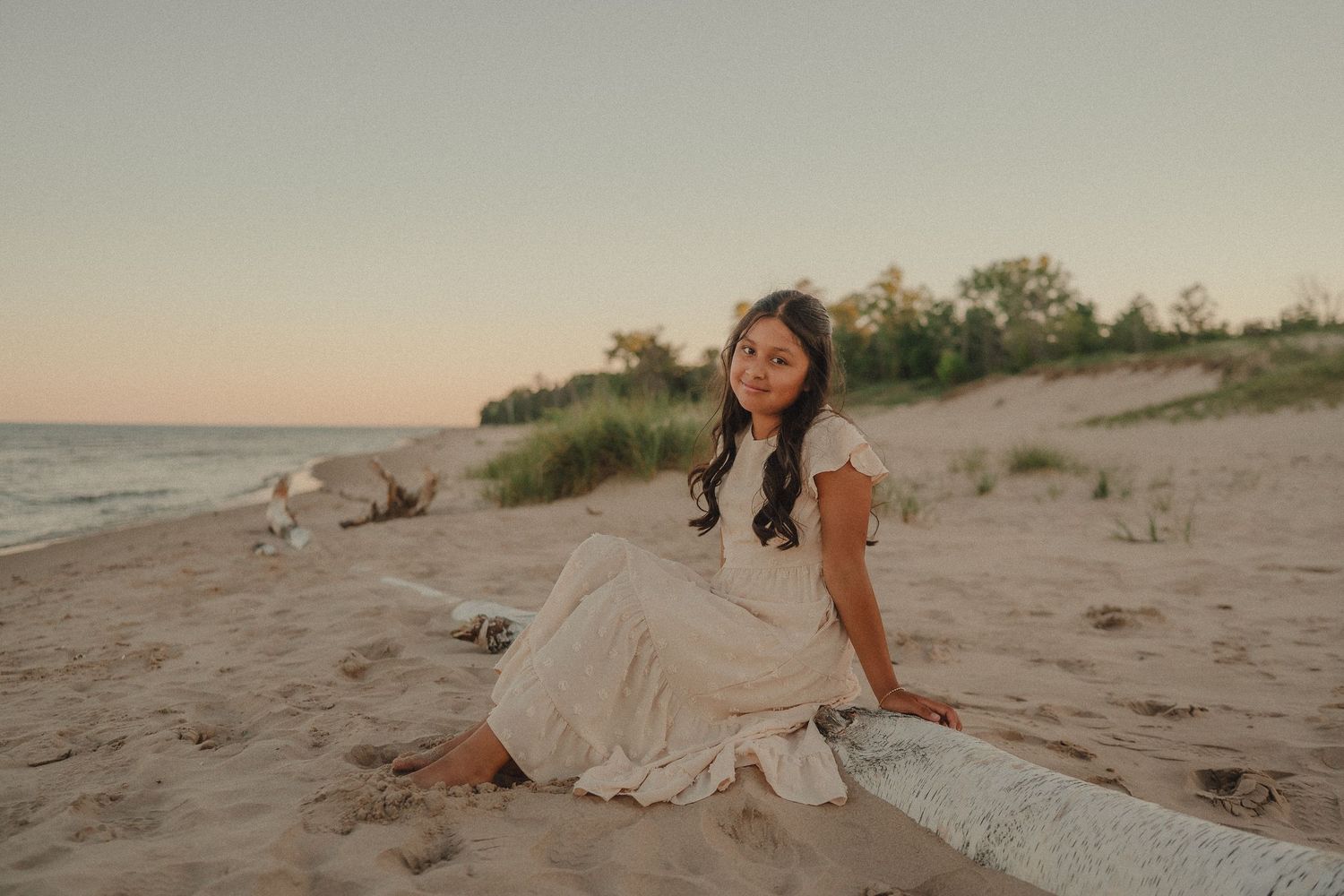 Young woman in cream dress sitting on driftwood on sandy beach at sunset.