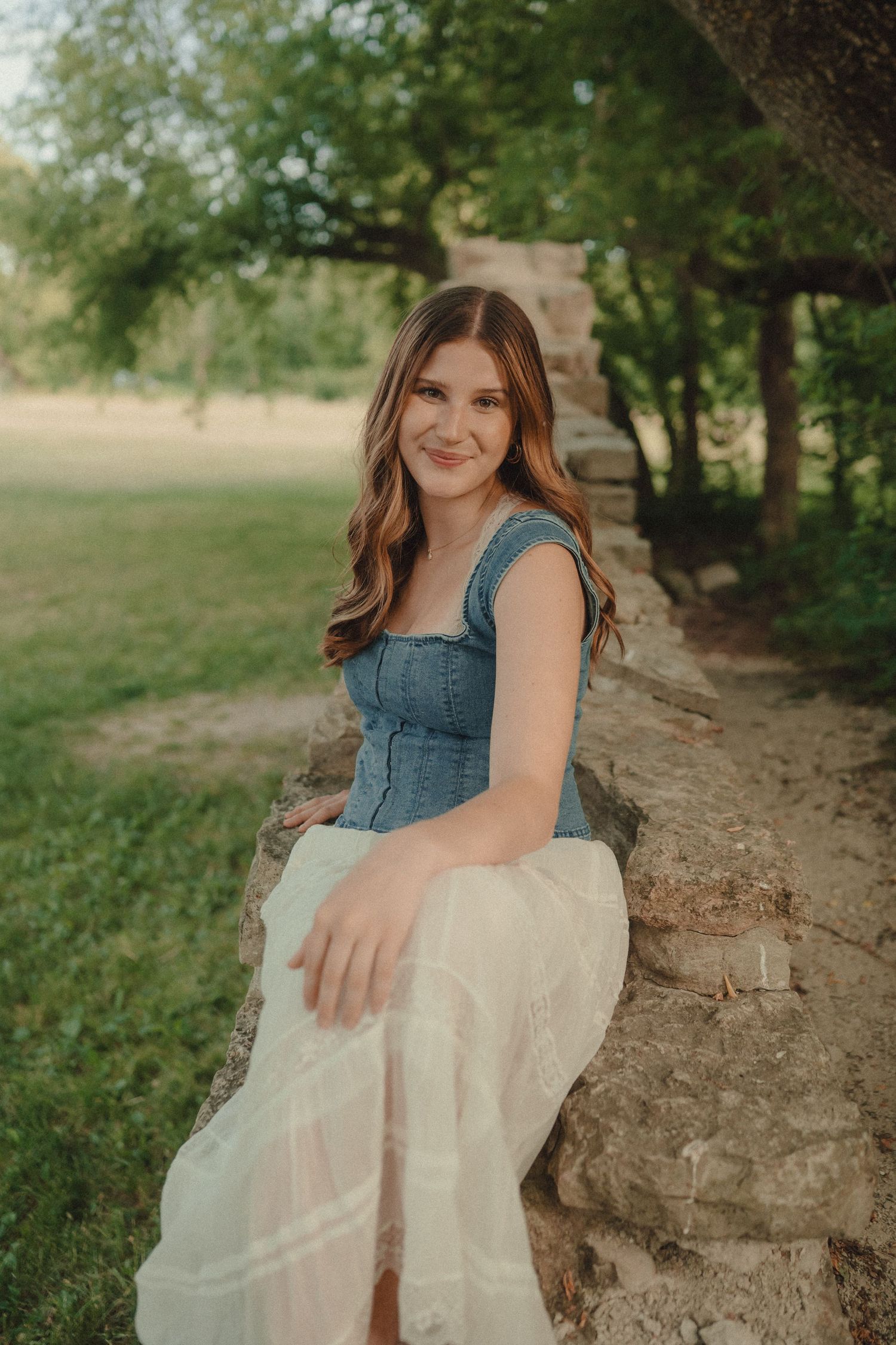 Young woman in denim top and white skirt sitting on stone wall in park.
