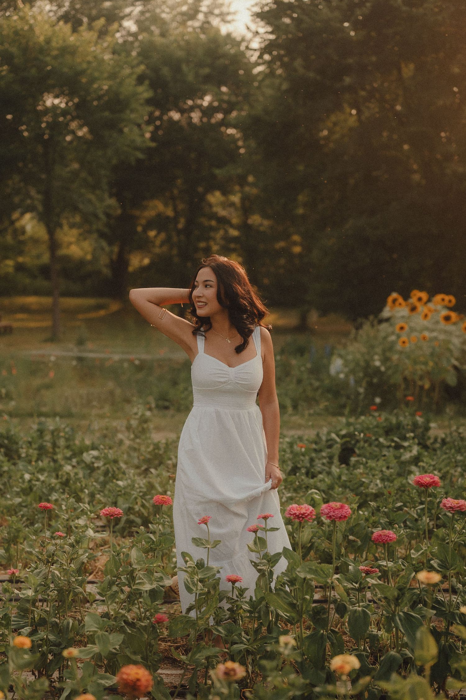 Woman in white sundress standing in a colorful flower garden with sunflowers and trees at golden hour.