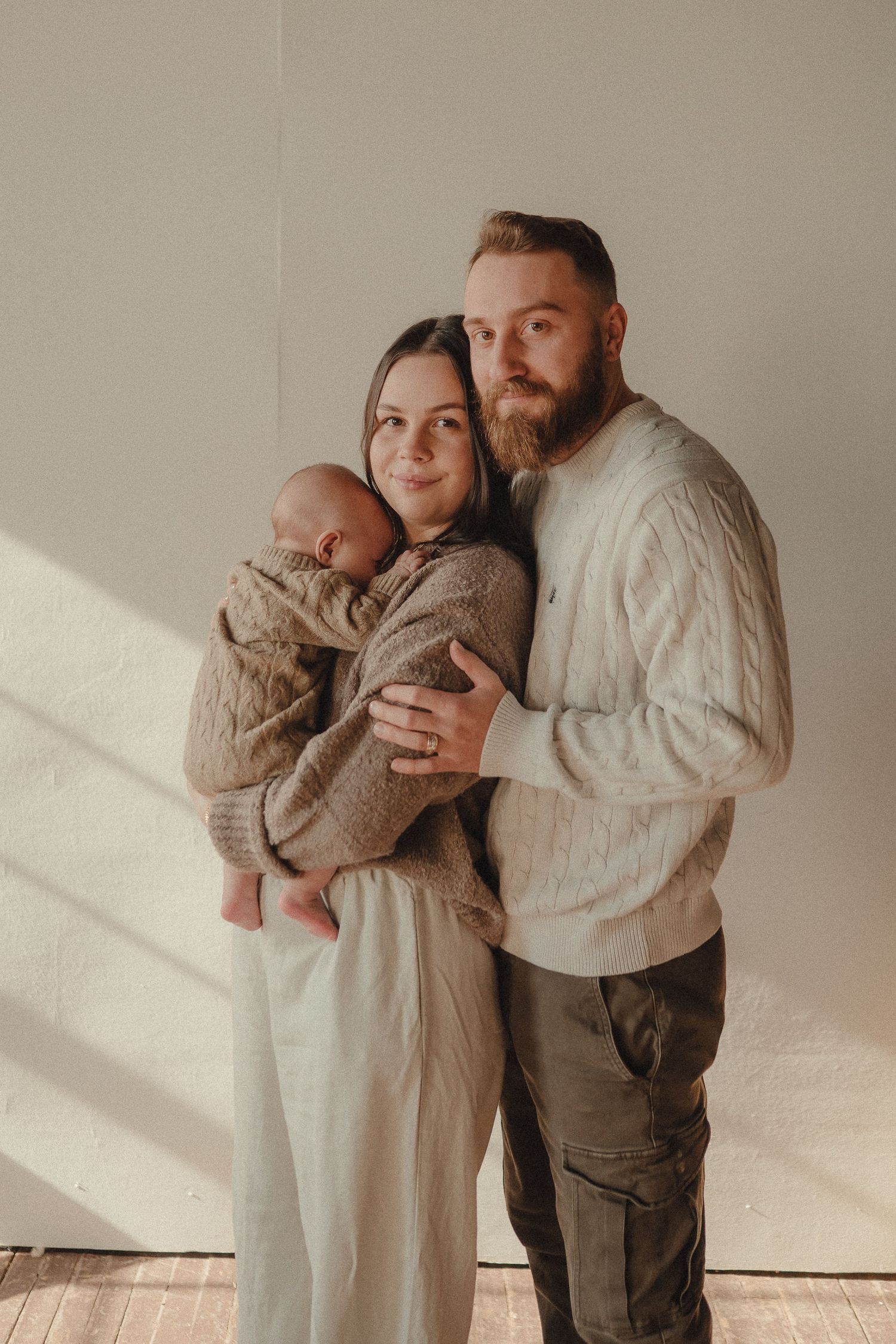 A family poses together in soft natural light wearing neutral colored clothing against a white wall.