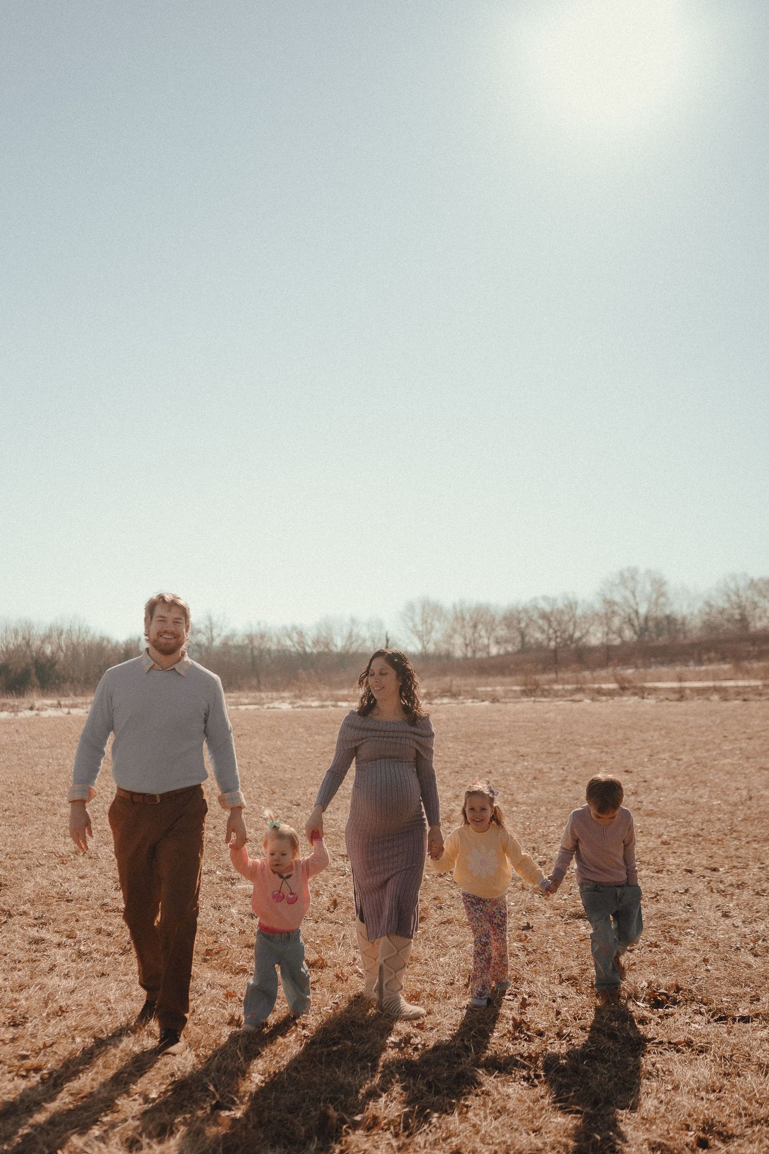 A family walks together holding hands across a sunlit field casting long shadows on the ground.