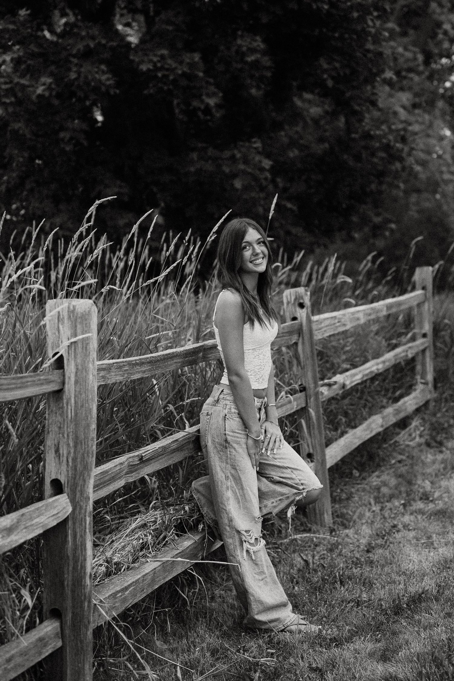 Black and white photo of smiling teenage girl leaning on wooden fence in field wearing jeans.