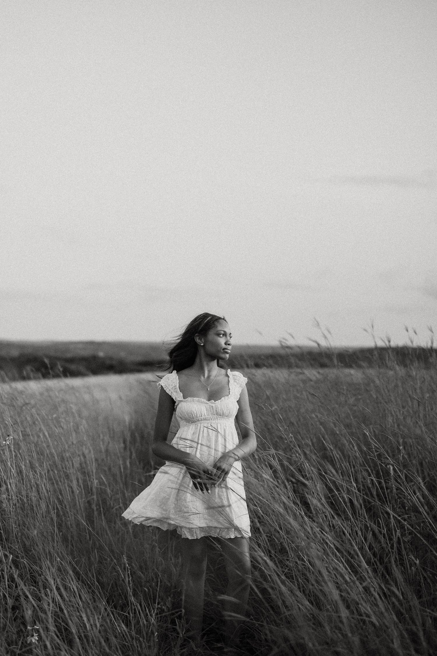 Black and white portrait of woman in white dress standing in tall grass field.