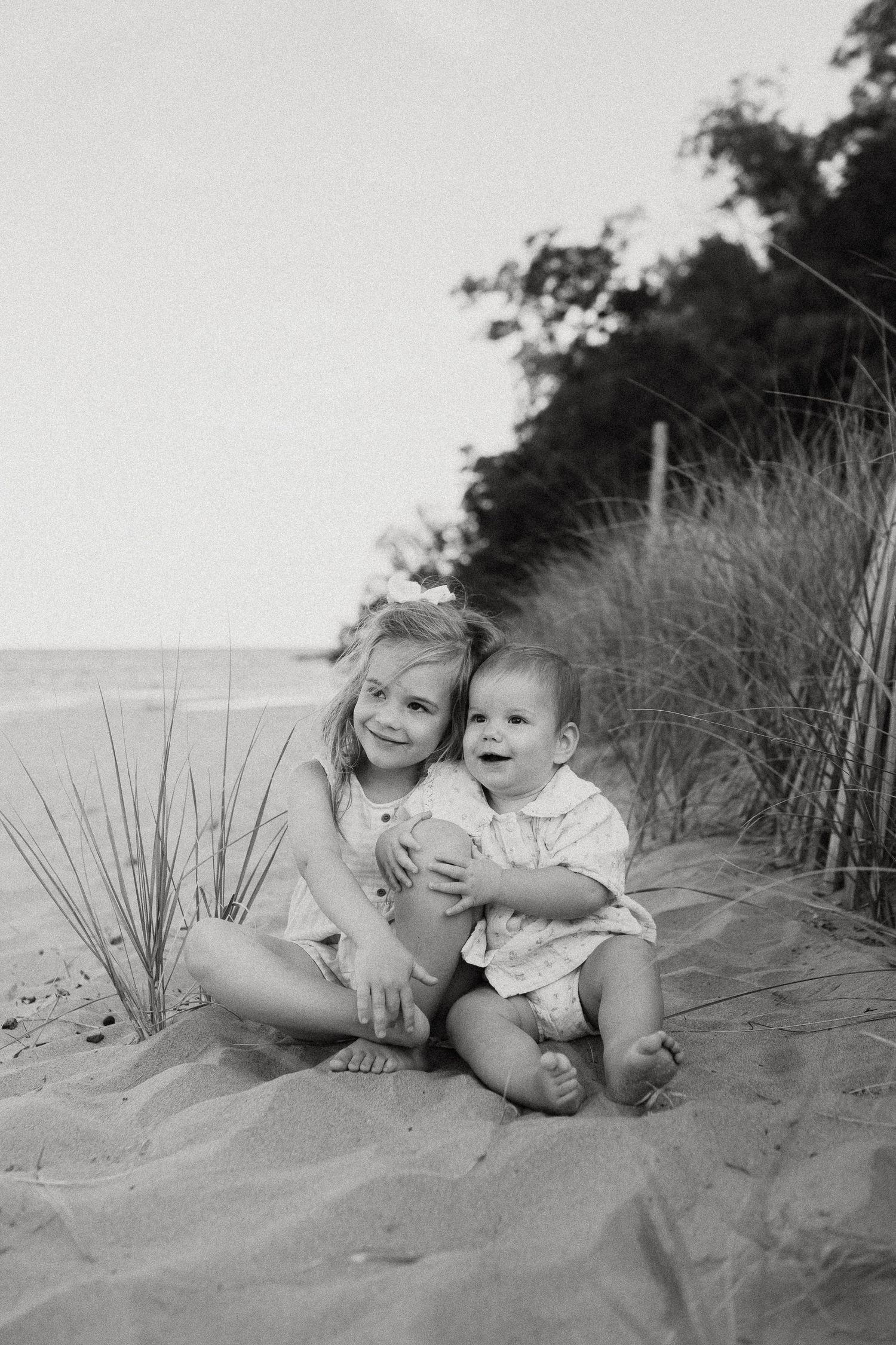 Black and white photo of two young siblings sitting together on sandy beach dunes, smiling joyfully.
